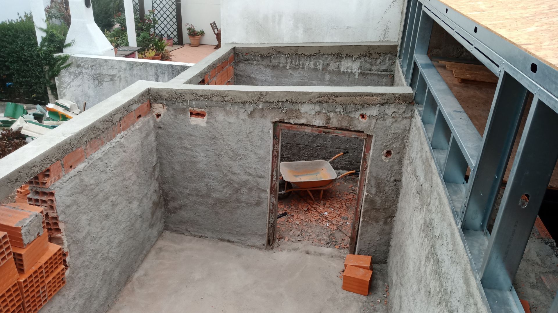 Canteiro de obras com paredes de concreto, alvenaria e estrutura metálica. Um carrinho de mão está dentro de uma porta.