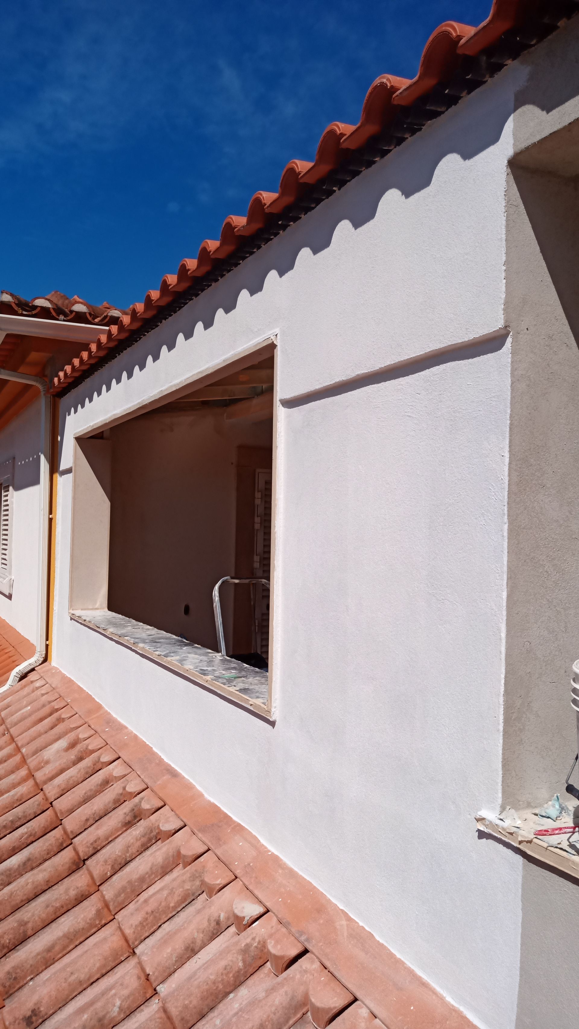 Exterior do edifício com uma janela; parede de estuque, telhado de telha vermelha, céu azul claro.