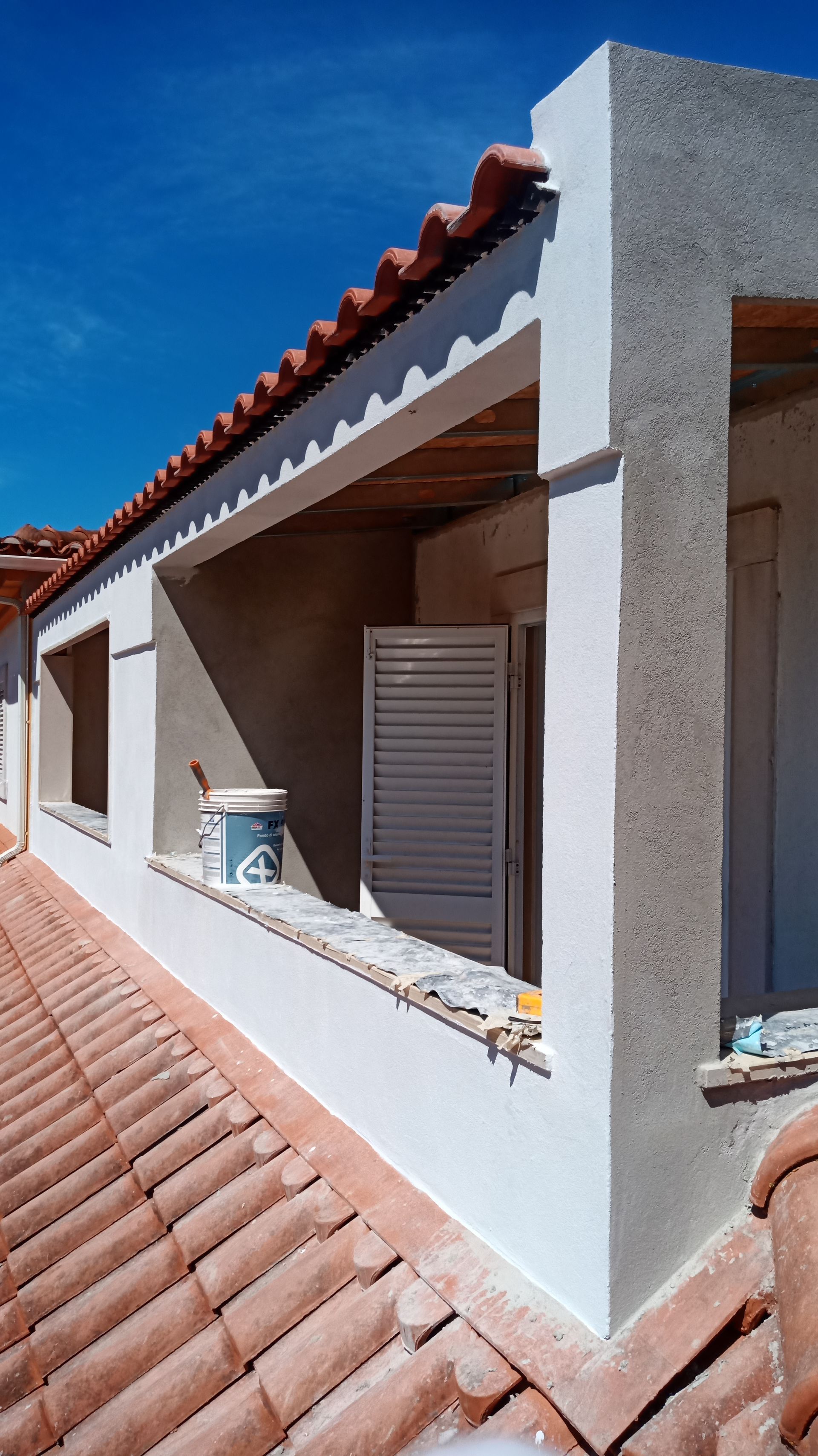 Exterior de um edifício com paredes brancas, telhado de telha vermelha e janela aberta com persianas, contra um céu azul.