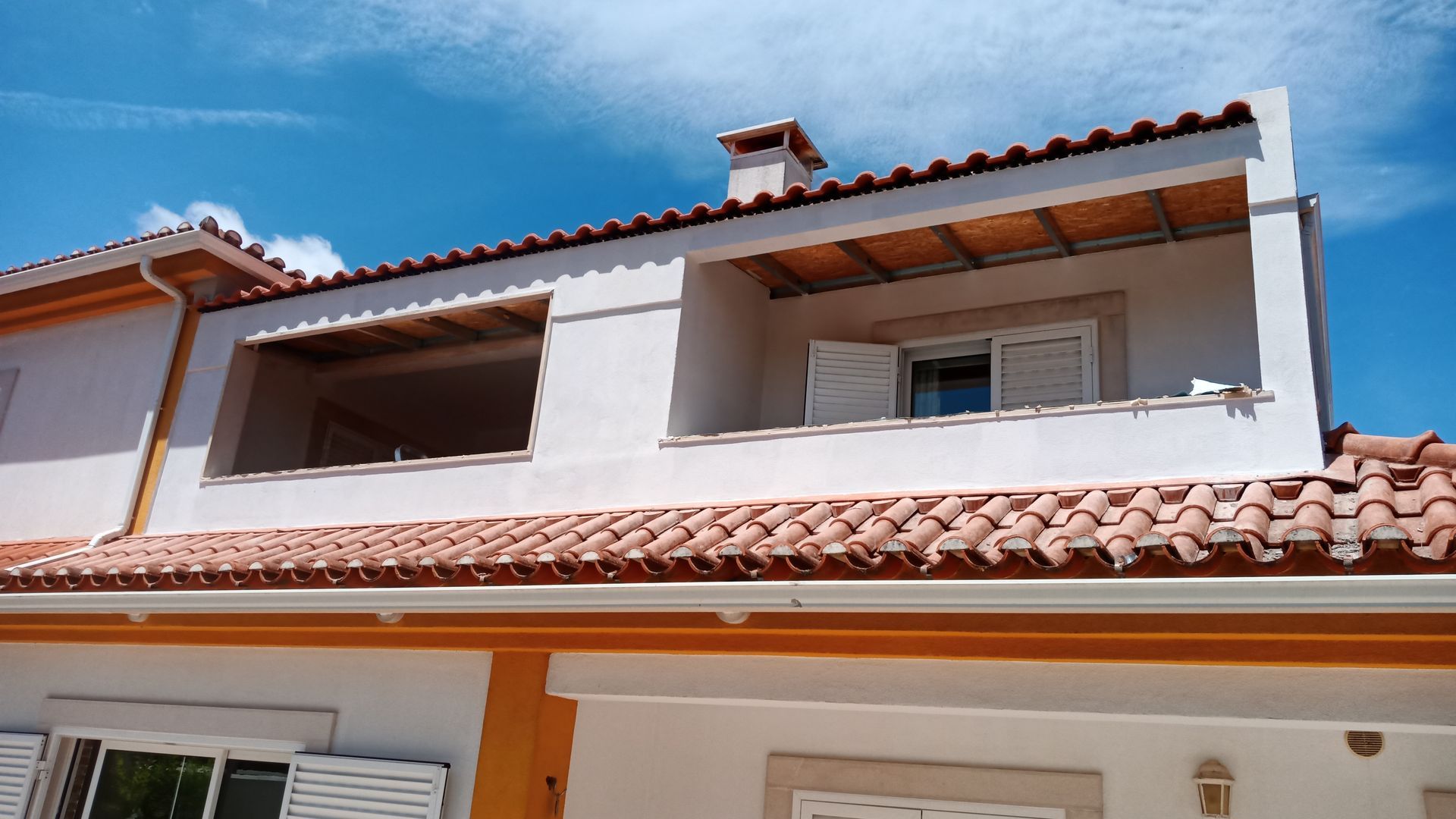 Prédio branco com telhado de telhas laranja e área de varanda externa sob um céu azul brilhante.