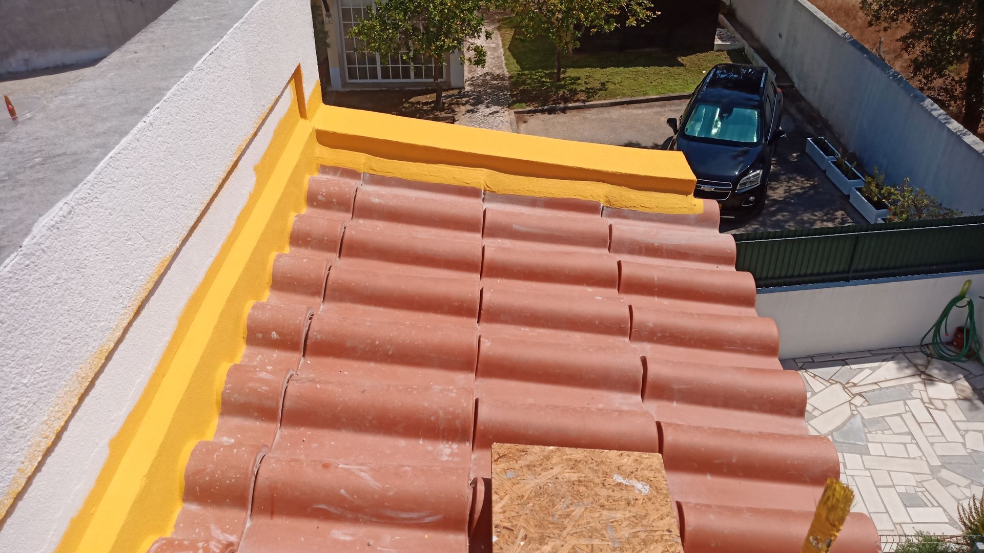 Telhado em construção: telhas vermelhas, isolamento amarelo e uma tábua de madeira visíveis. Um carro e árvores ao fundo.