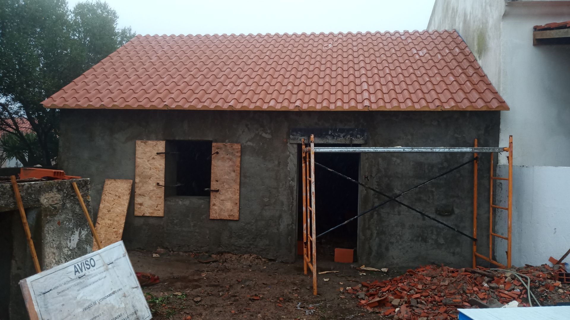 Uma pequena construção de concreto com telhado de telha vermelha; tábuas de madeira sobre uma janela; andaimes.