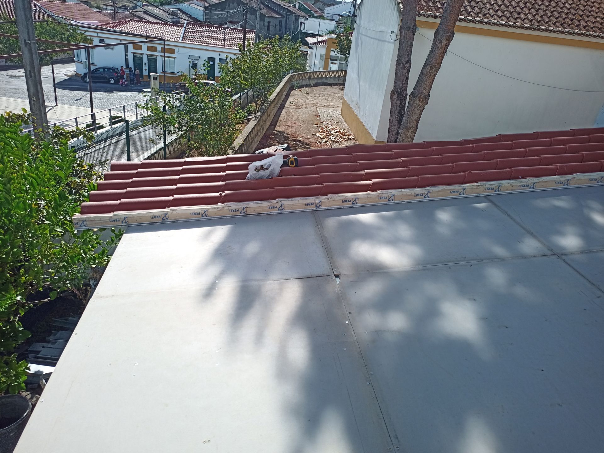 Vista aérea de um telhado com telhas vermelhas, cobertura metálica branca e um fundo de edifícios e vegetação.