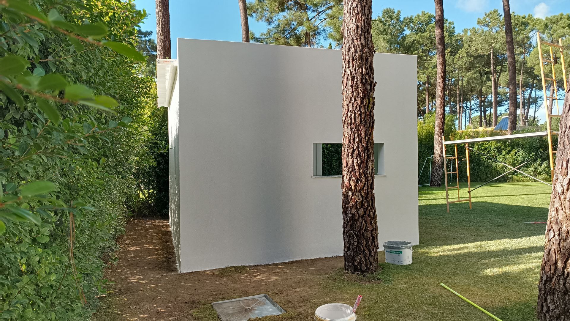 Prédio quadrado branco com uma pequena janela, rodeado por árvores e vegetação.