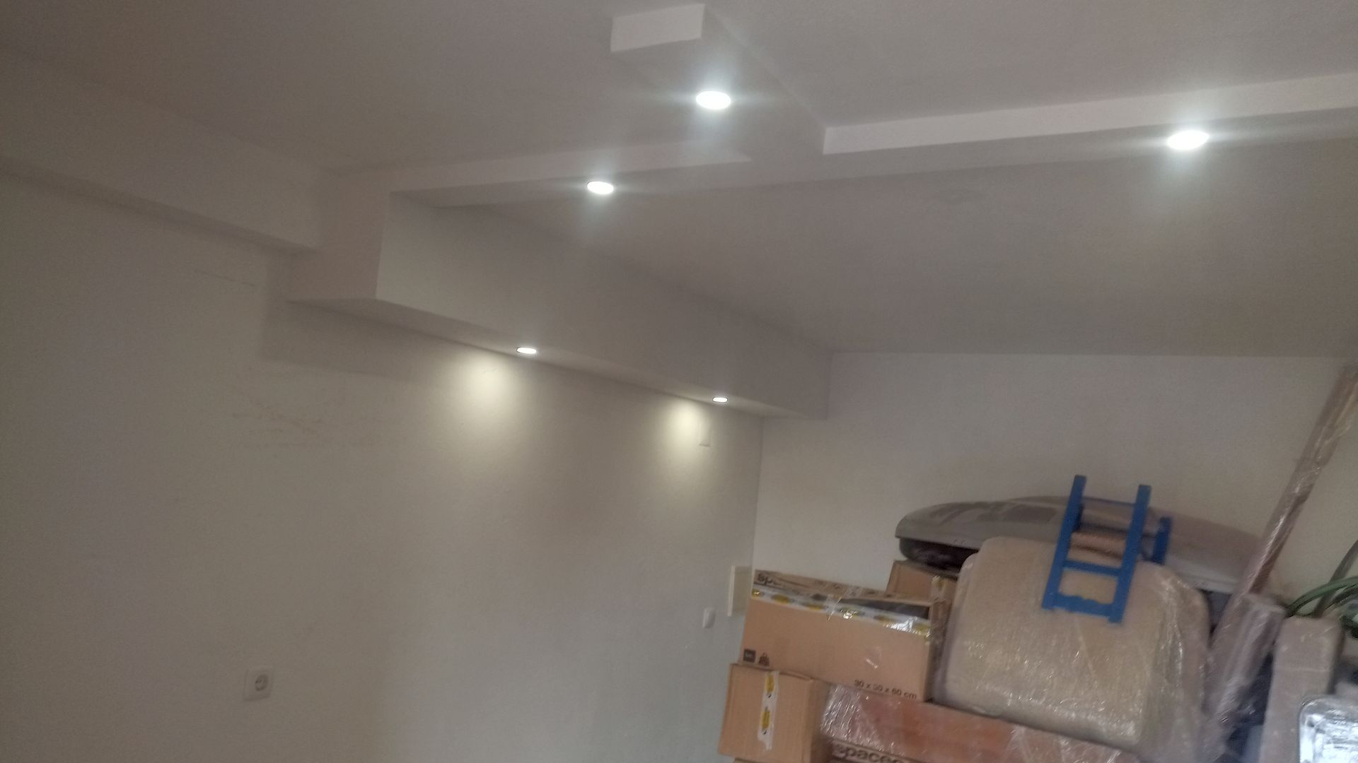 Vista interna de um cômodo com iluminação embutida em uma seção do teto e da parede. Caixas e entulhos estão empilhados no chão.