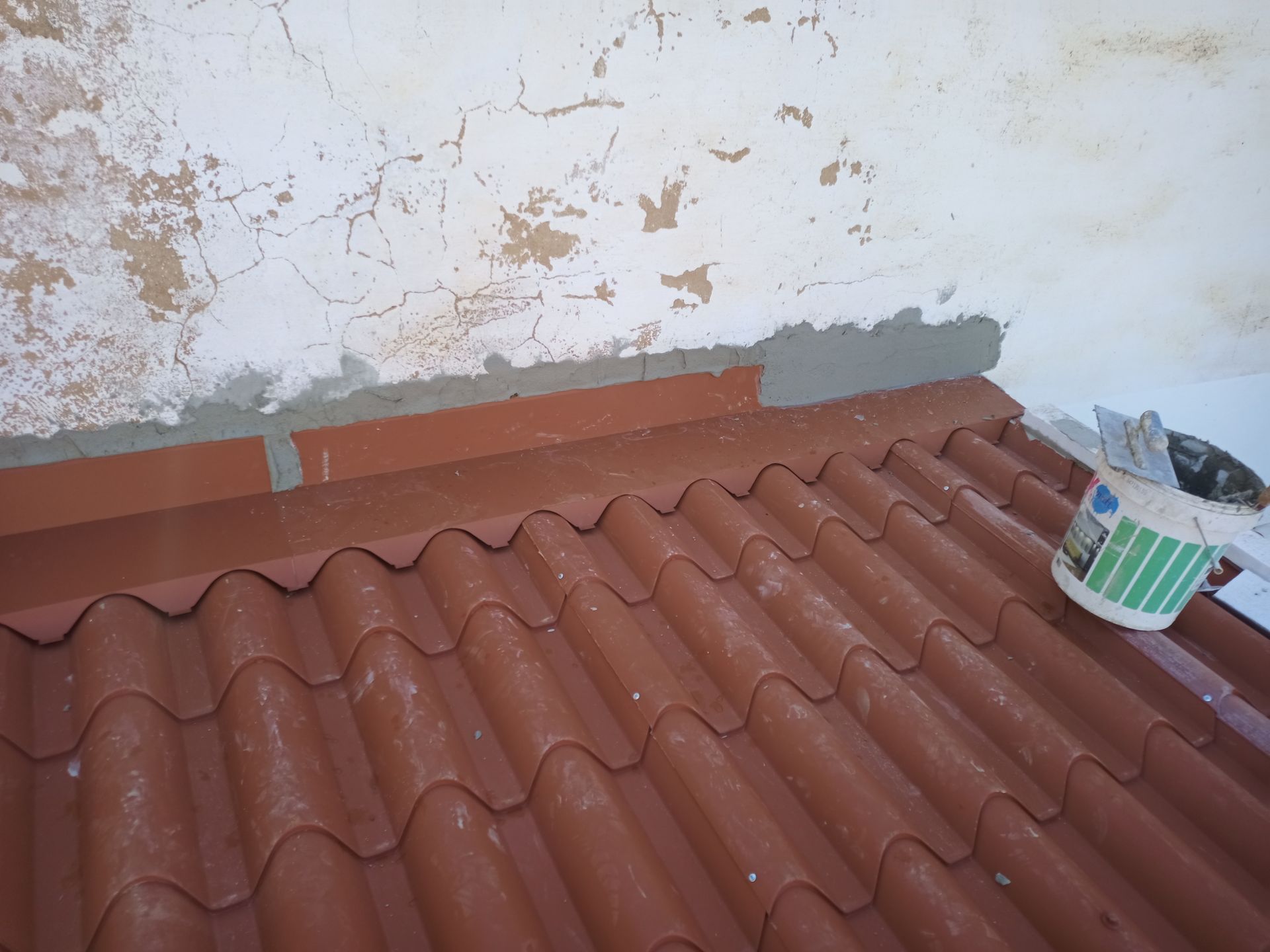 Telhado de telhas vermelhas contra uma parede branca desgastada, com argamassa e um balde de tinta visíveis.