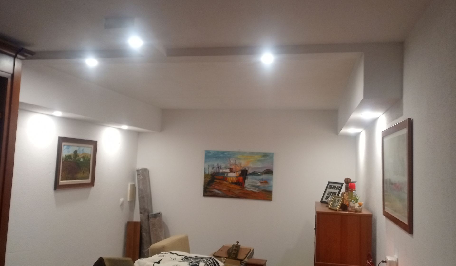 Um quarto com paredes brancas, luzes embutidas no teto e obras de arte. Um armário de madeira e alguns móveis são visíveis.