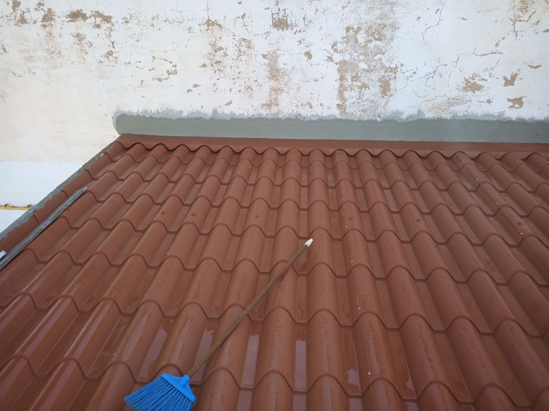 Telhado de telha marrom com uma faixa cinza contra uma parede branca com tinta descascando; uma vassoura azul repousa sobre o telhado.