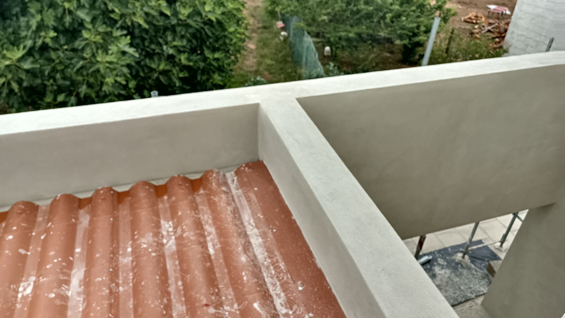 Detalhe de um telhado ondulado marrom sob uma estrutura de concreto bege, com vista para a folhagem verde ao fundo.