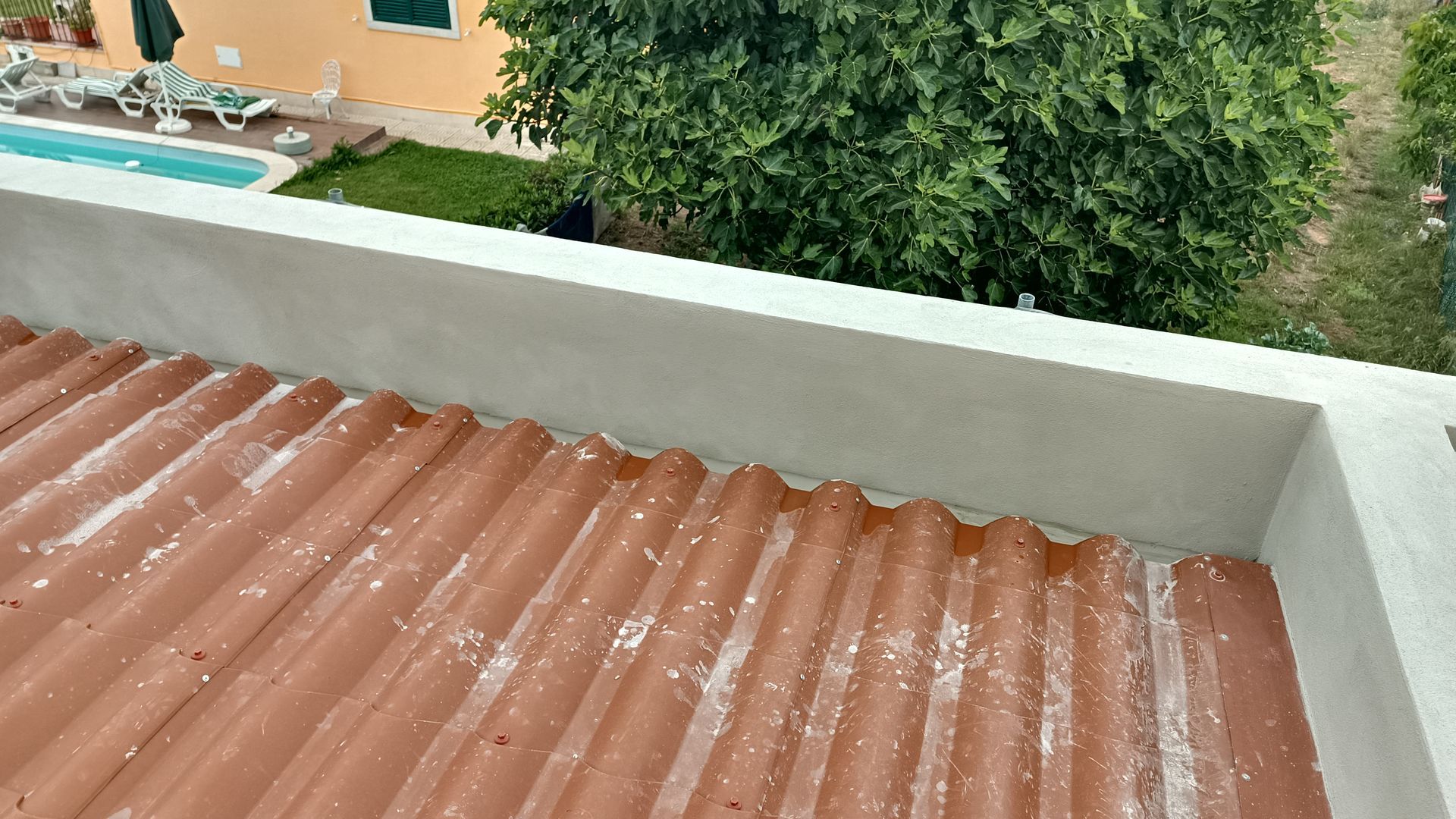 Telhado ondulado avermelhado com parede branca e vegetação ao fundo, provavelmente em ambiente externo.