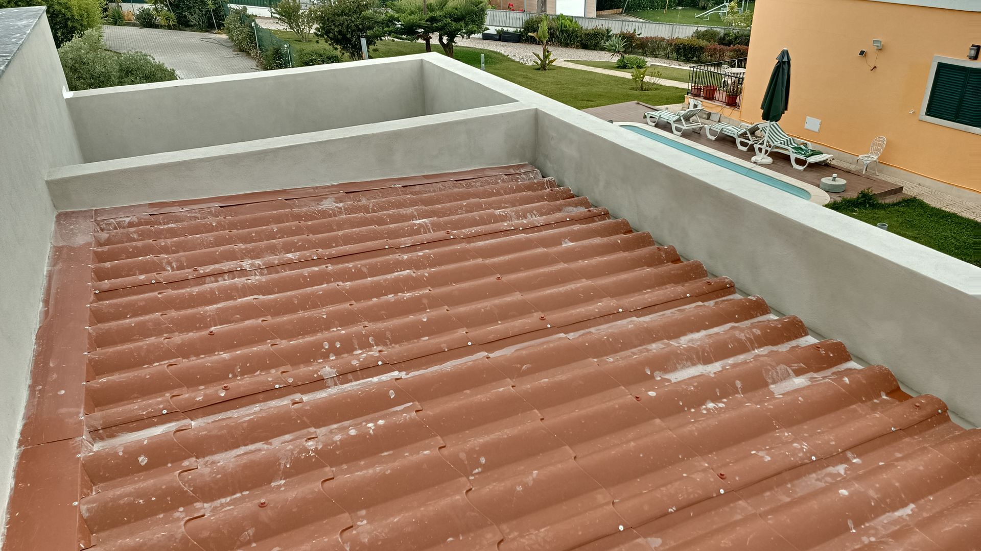 Telhado de zinco vermelho com parede de concreto claro, cercado por área residencial, com piscina visível.