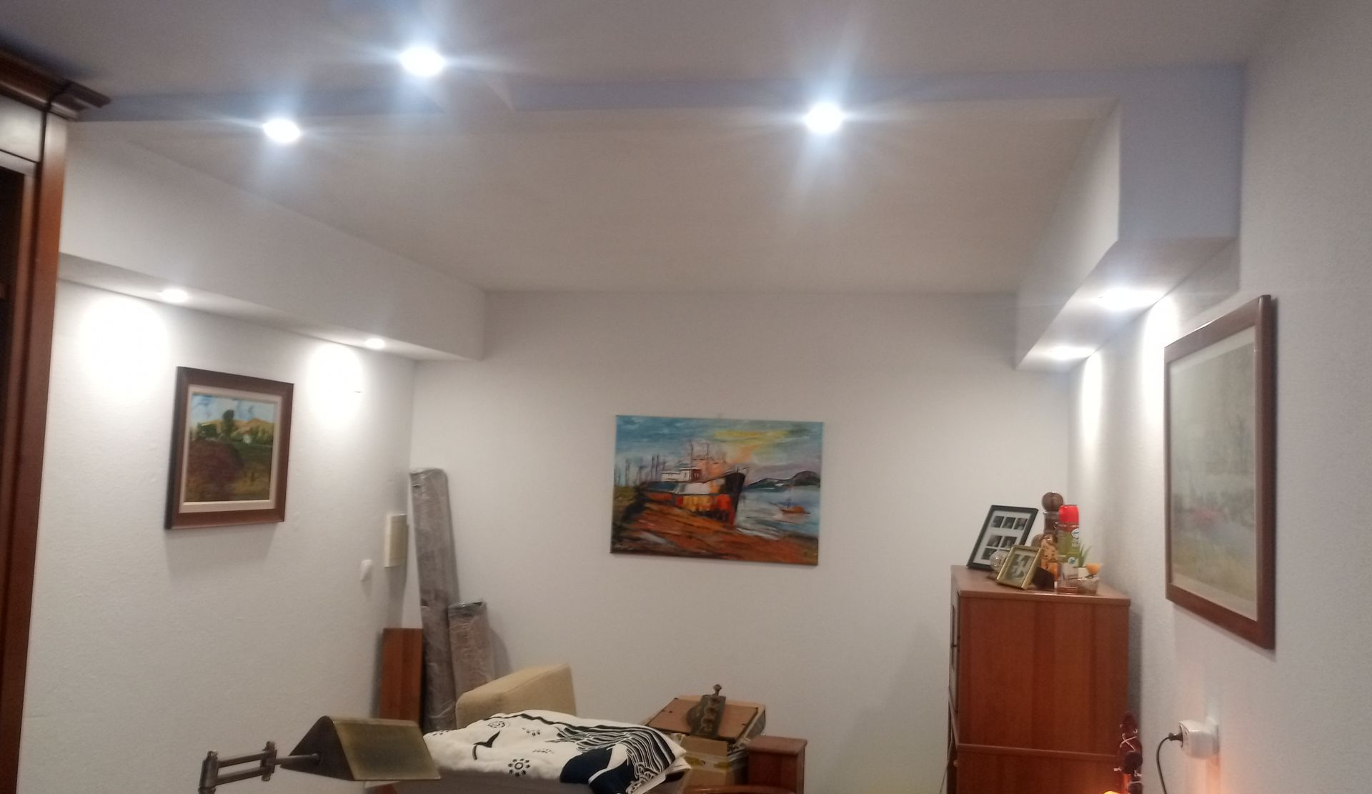 Quarto com paredes brancas e luzes embutidas no teto; obras de arte nas paredes; móveis e objetos espalhados.