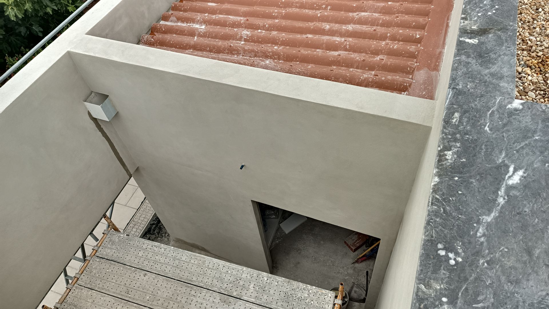 Escadaria que desce para uma pequena sala fechada com uma porta. Telhado de zinco vermelho visível acima. Construção em concreto.