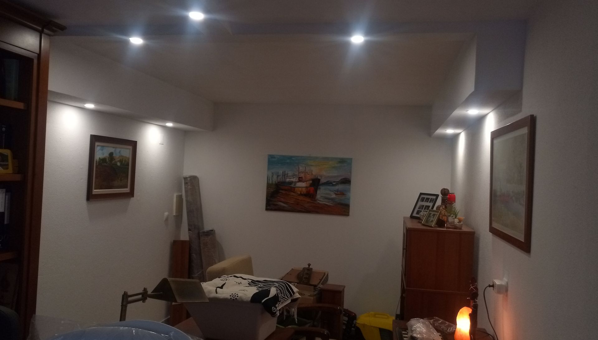 Quarto aconchegante com paredes brancas, iluminação embutida e obras de arte. Um armário de madeira e diversos objetos estão em primeiro plano.