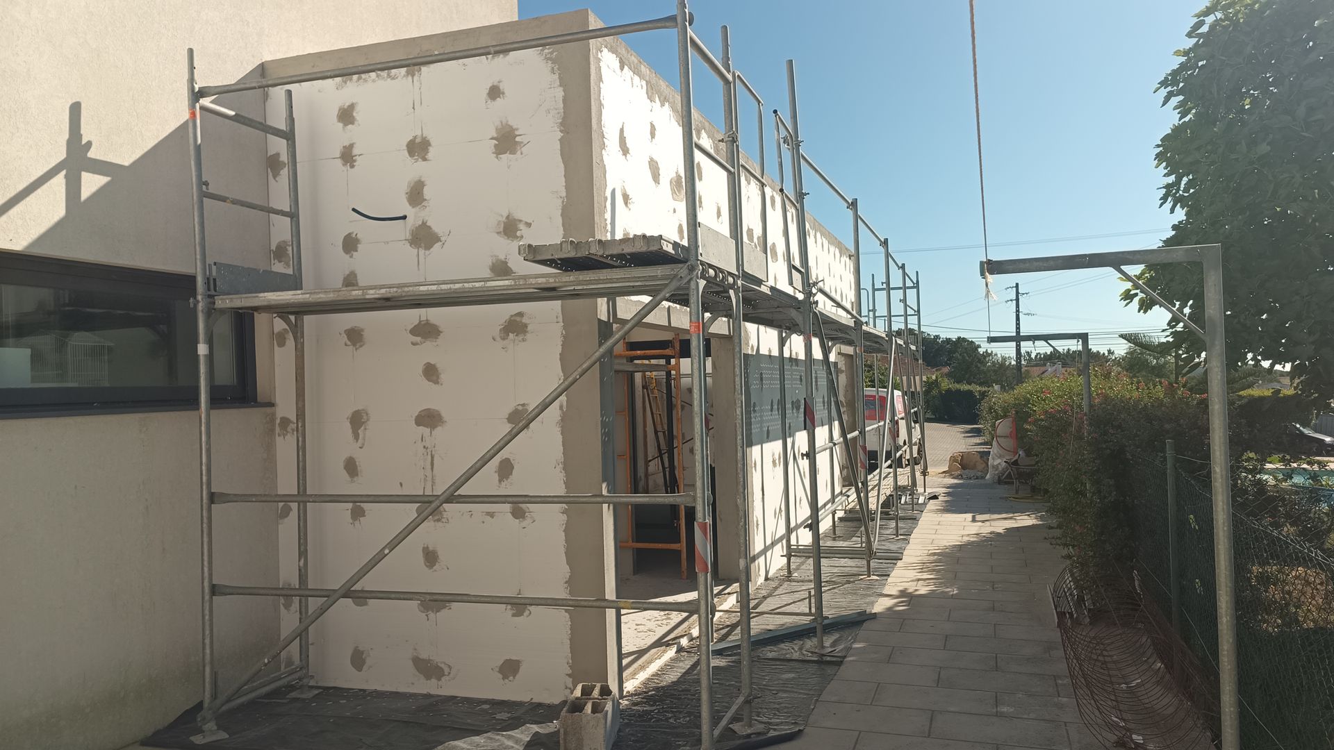 Exterior de um edifício em construção; andaimes envolvem uma estrutura com isolamento exposto, ao lado de uma passagem.