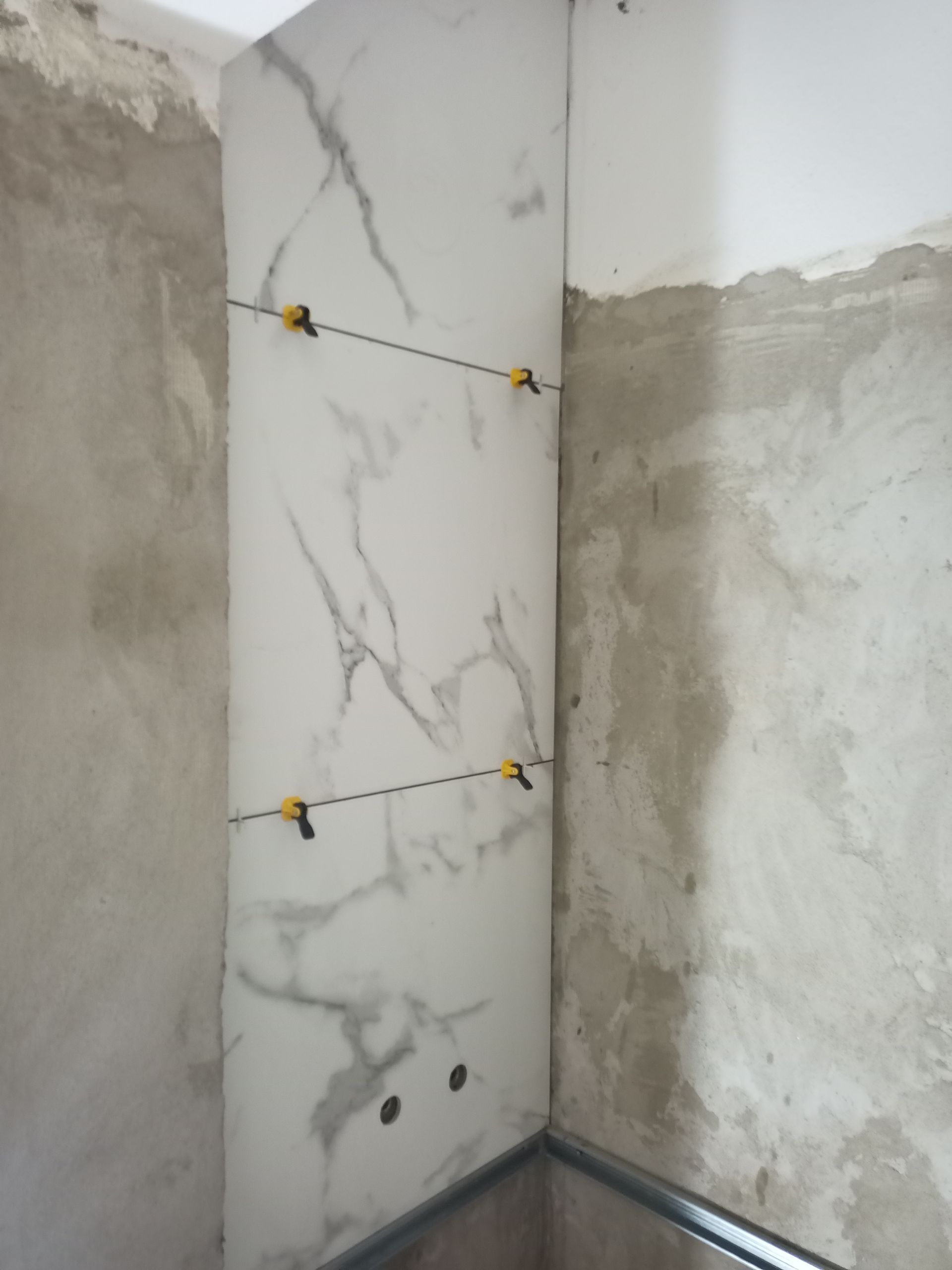 Parede de canto revestida com azulejos brancos com veios cinzentos, com grampos de nivelamento visíveis. Ambiente de construção.