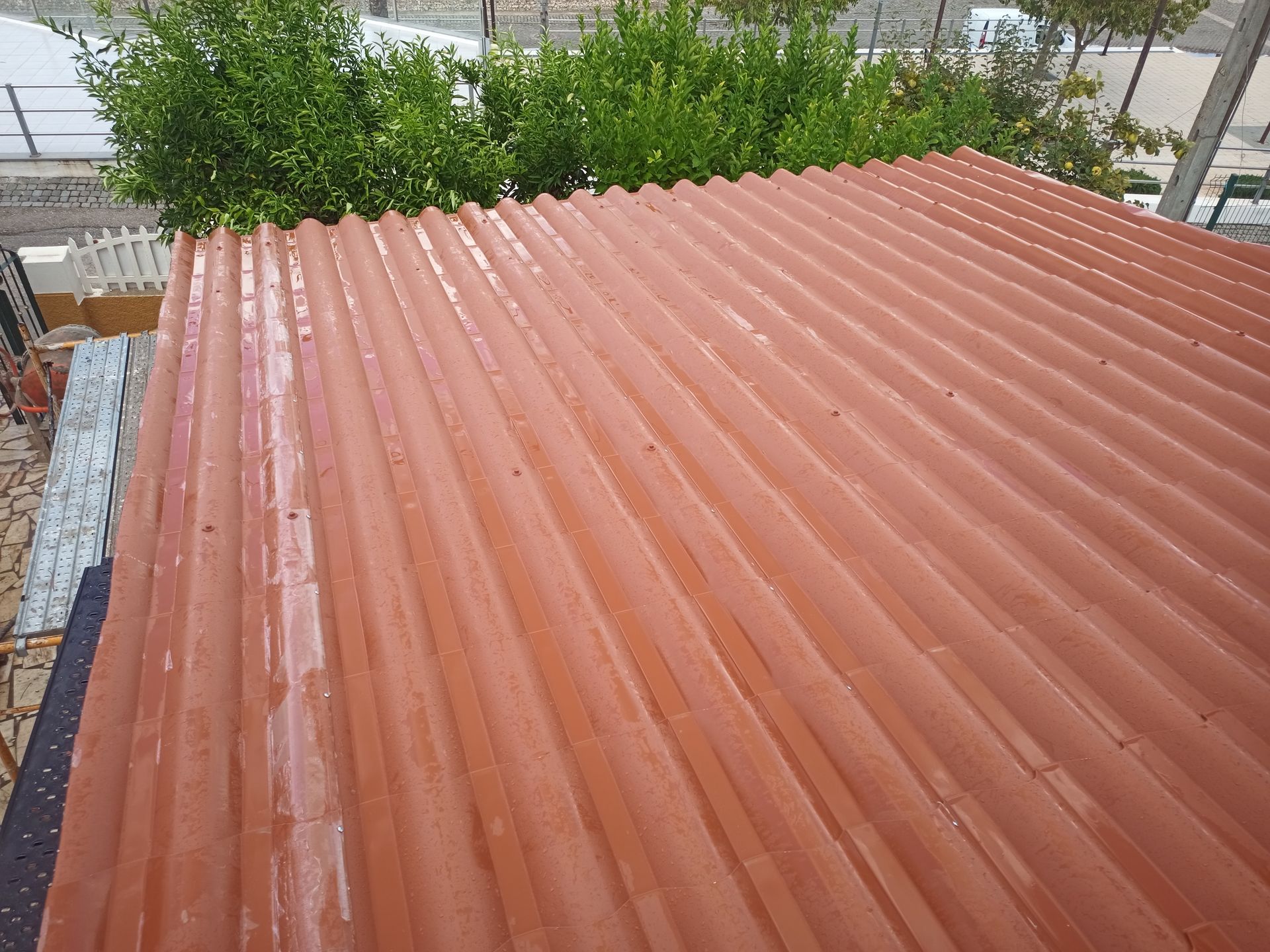 Telhado de metal ondulado vermelho, molhado pela chuva, com folhagem verde ao fundo.