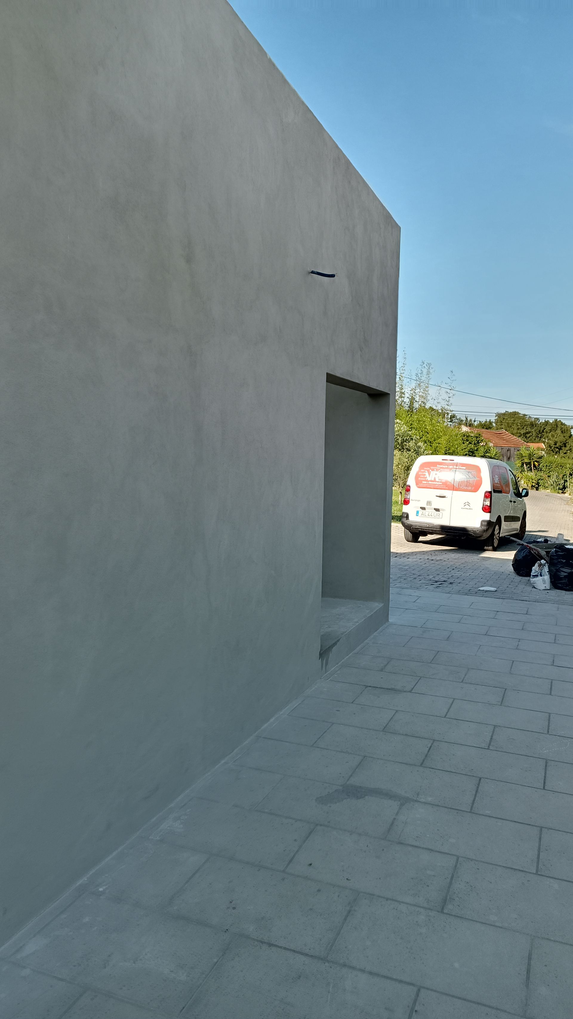 Parede rebocada em cinza com uma porta recuada. Uma van está estacionada nas proximidades sob um céu azul.