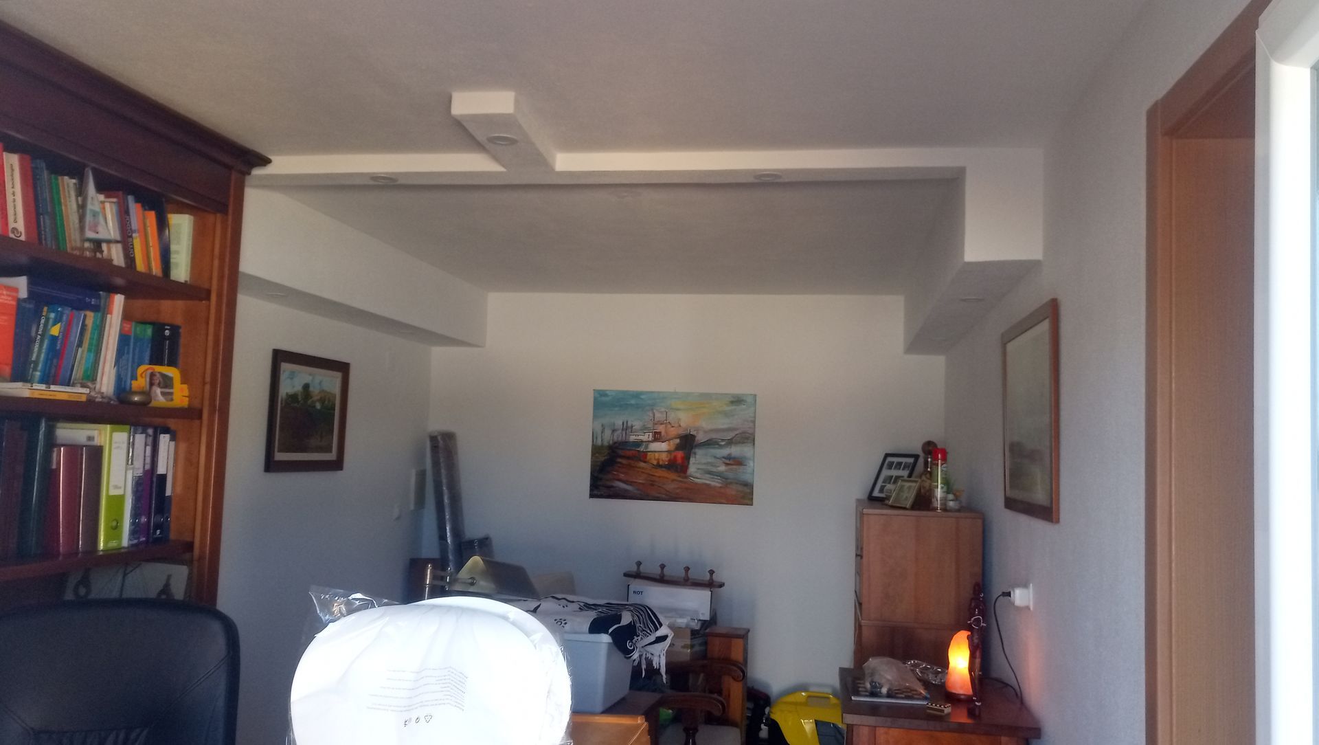 Quarto com estante de livros, obras de arte e escrivaninha desarrumada. Paredes brancas, móveis de madeira e iluminação no teto.