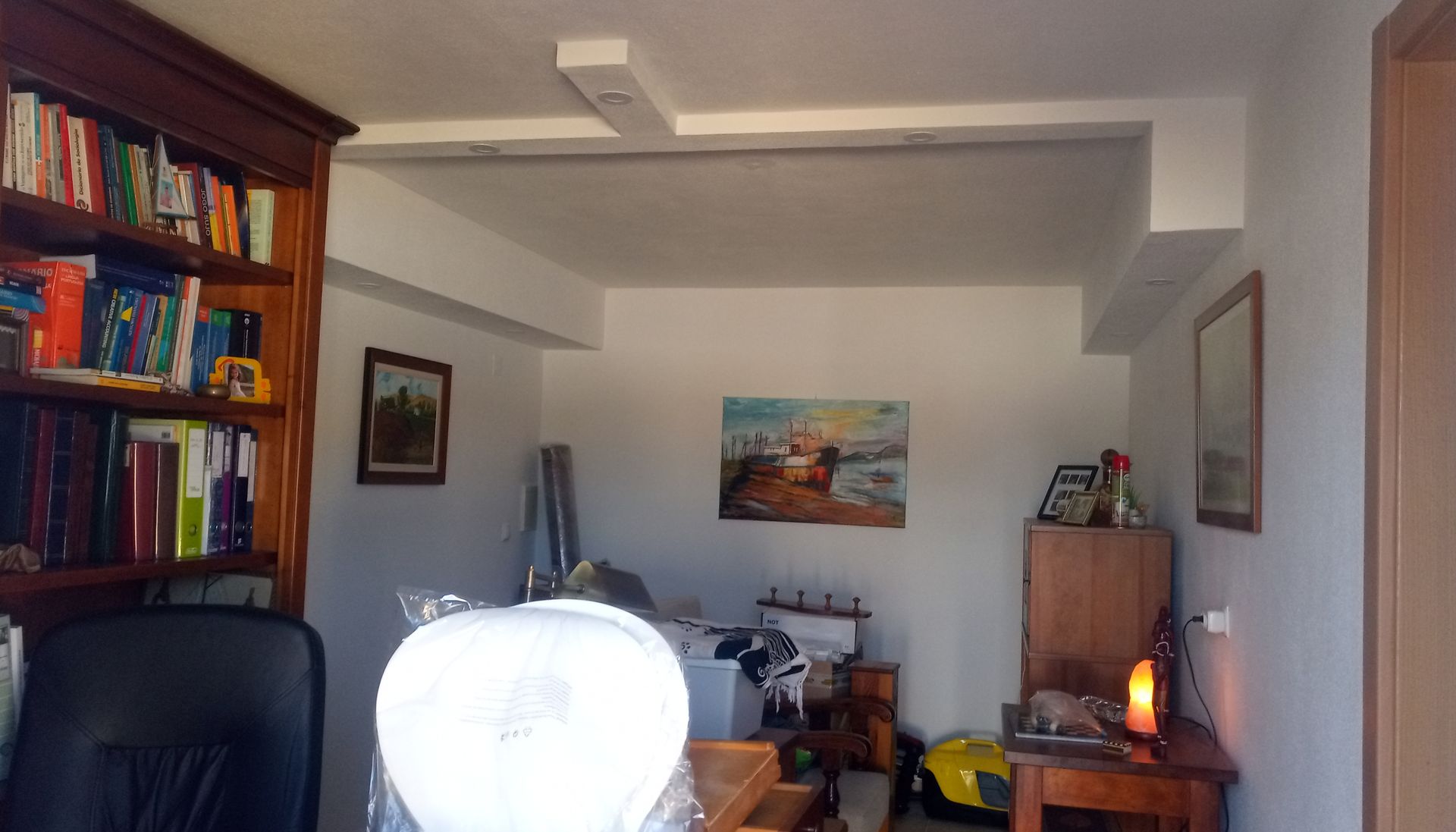 Um quarto com uma estante de livros, uma escrivaninha e obras de arte. Um ventilador e objetos diversos estão em primeiro plano.