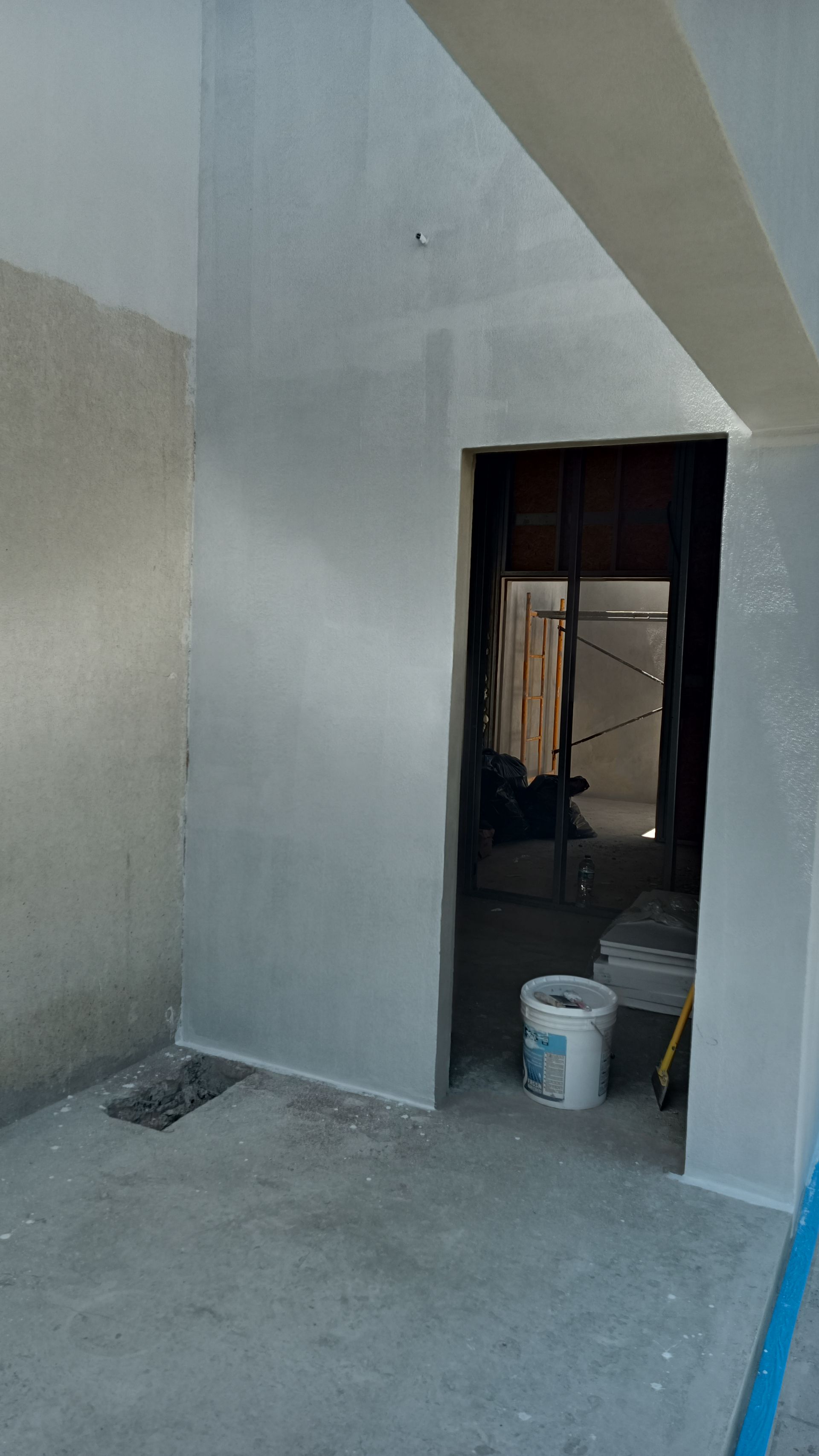 Quarto interior em construção, com porta aberta, paredes cinzentas e um balde branco.