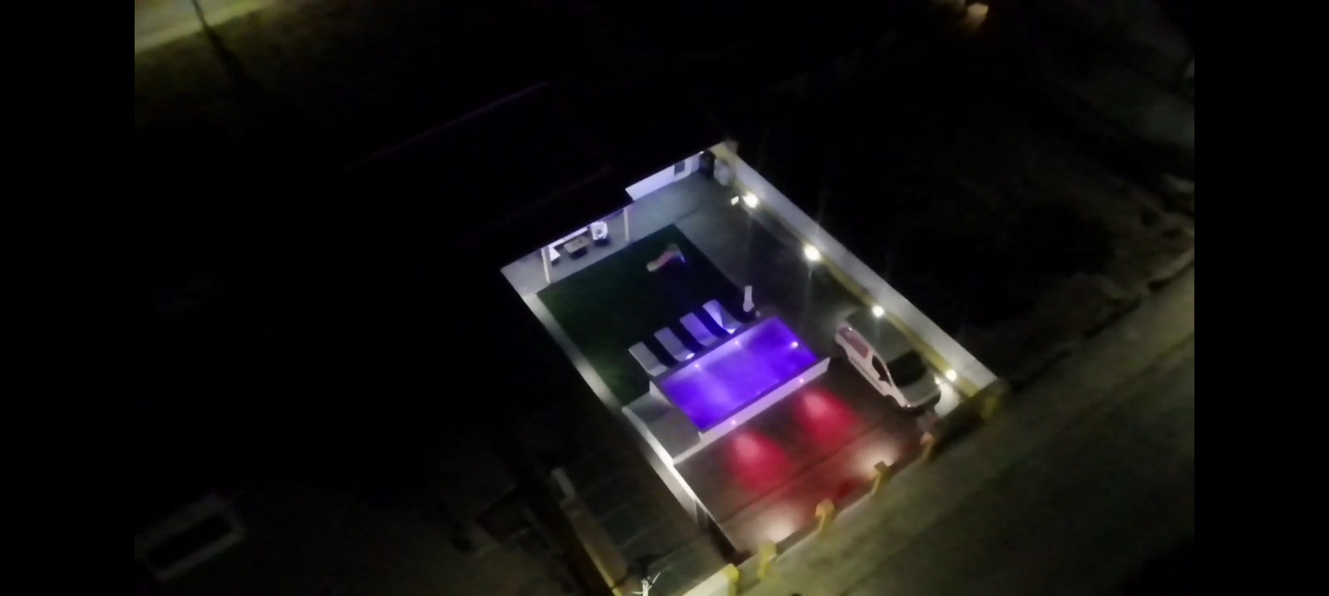 Vista aérea noturna de uma estrutura retangular bem iluminada com piscina e assentos.
