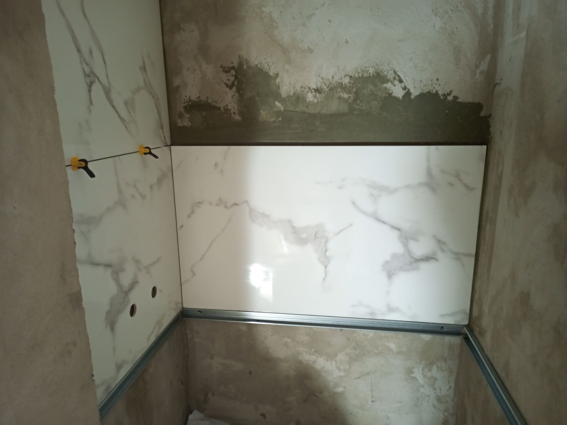 Instalação de azulejos na parede de um banheiro, com grampos de nivelamento e acabamento metálico.