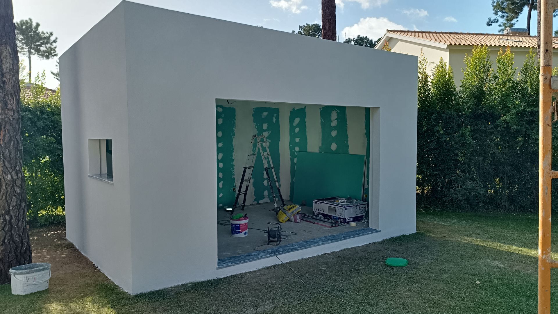 Um pequeno prédio branco em construção ao ar livre. É possível ver o gesso cartonado verde na entrada aberta.