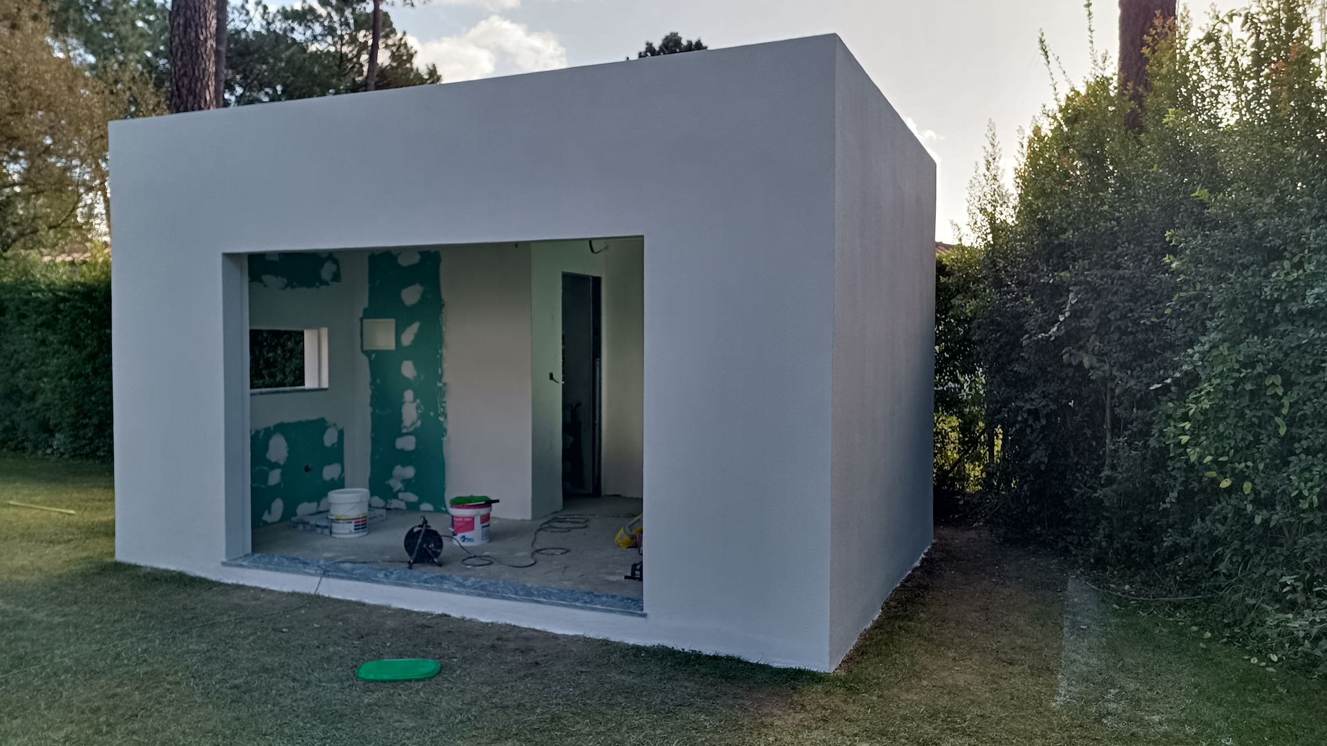 Prédio retangular branco, inacabado, com a fachada aberta em um gramado, com ferramentas de construção visíveis no interior.