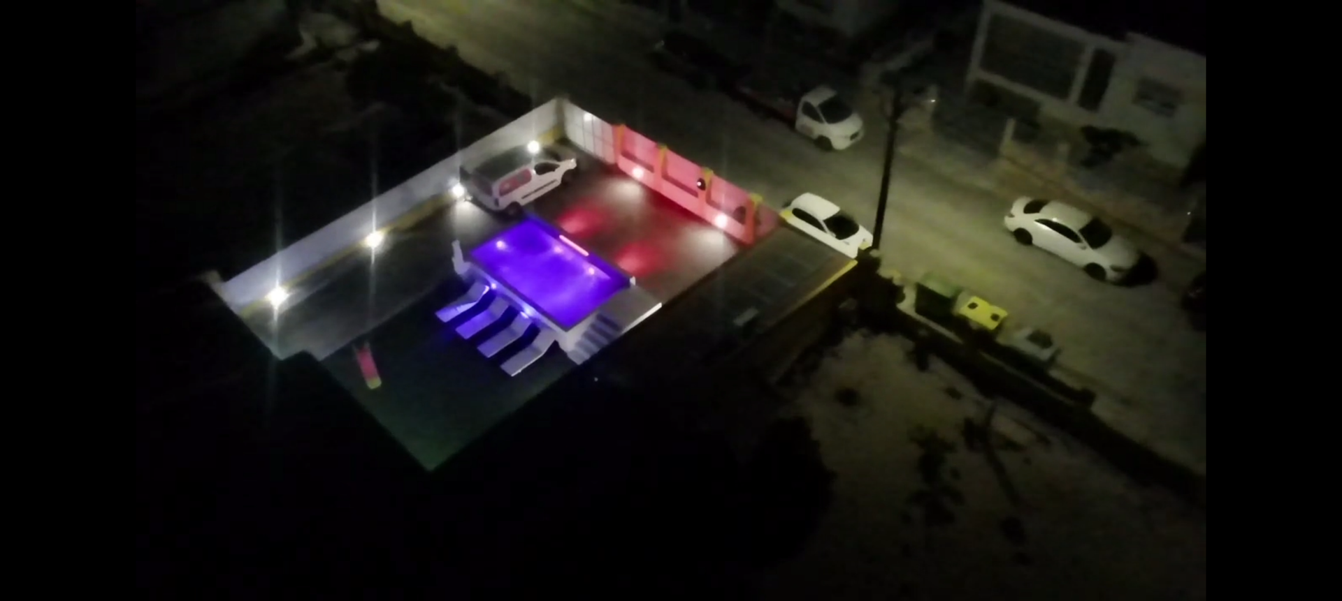 Vista aérea noturna de uma piscina com água roxa, espreguiçadeiras brancas, luminárias vermelhas na parede e rua com carros brancos.