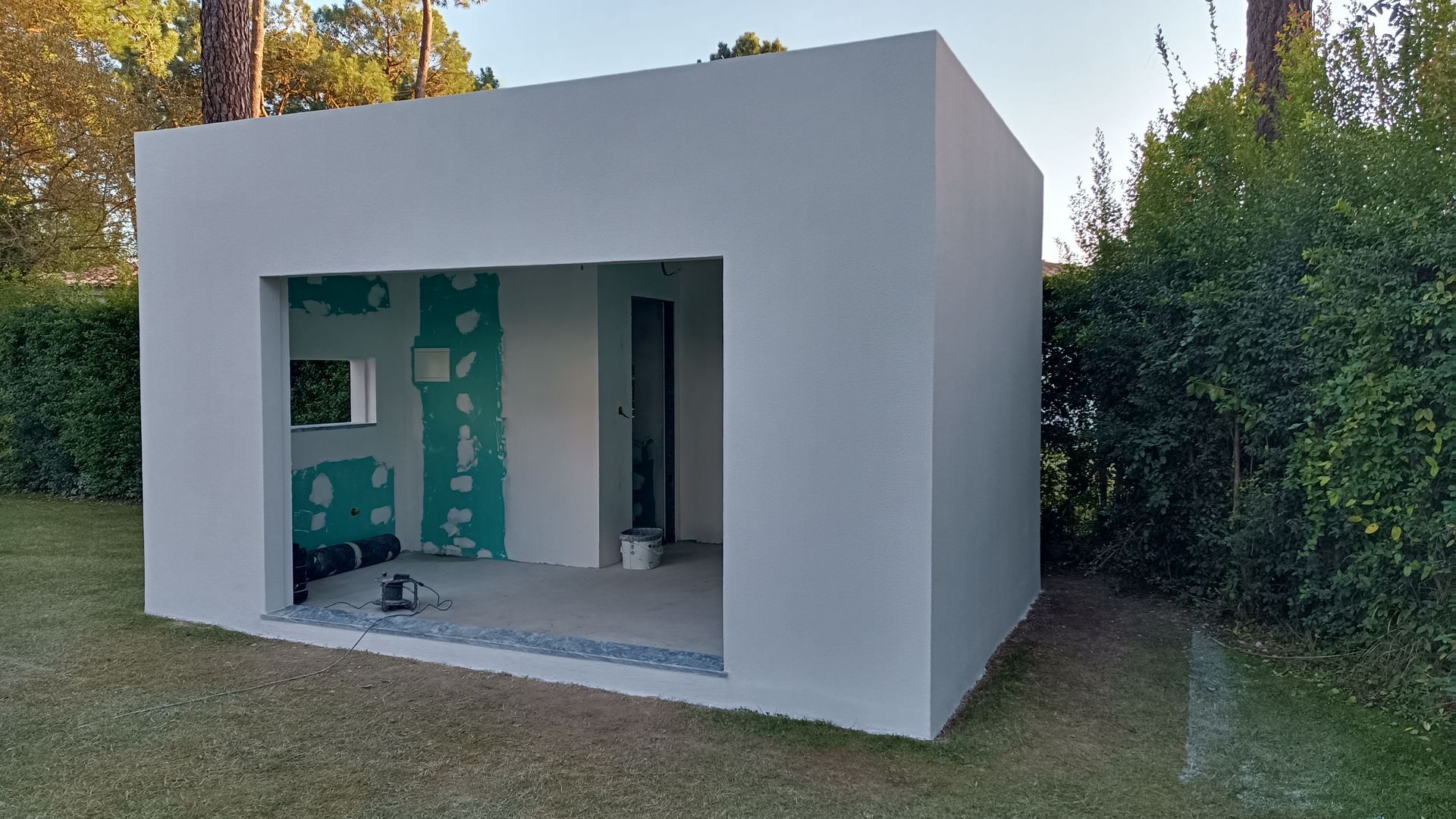 Prédio quadrado branco com entrada aberta em um gramado, parcialmente em construção, com paredes internas inacabadas visíveis.