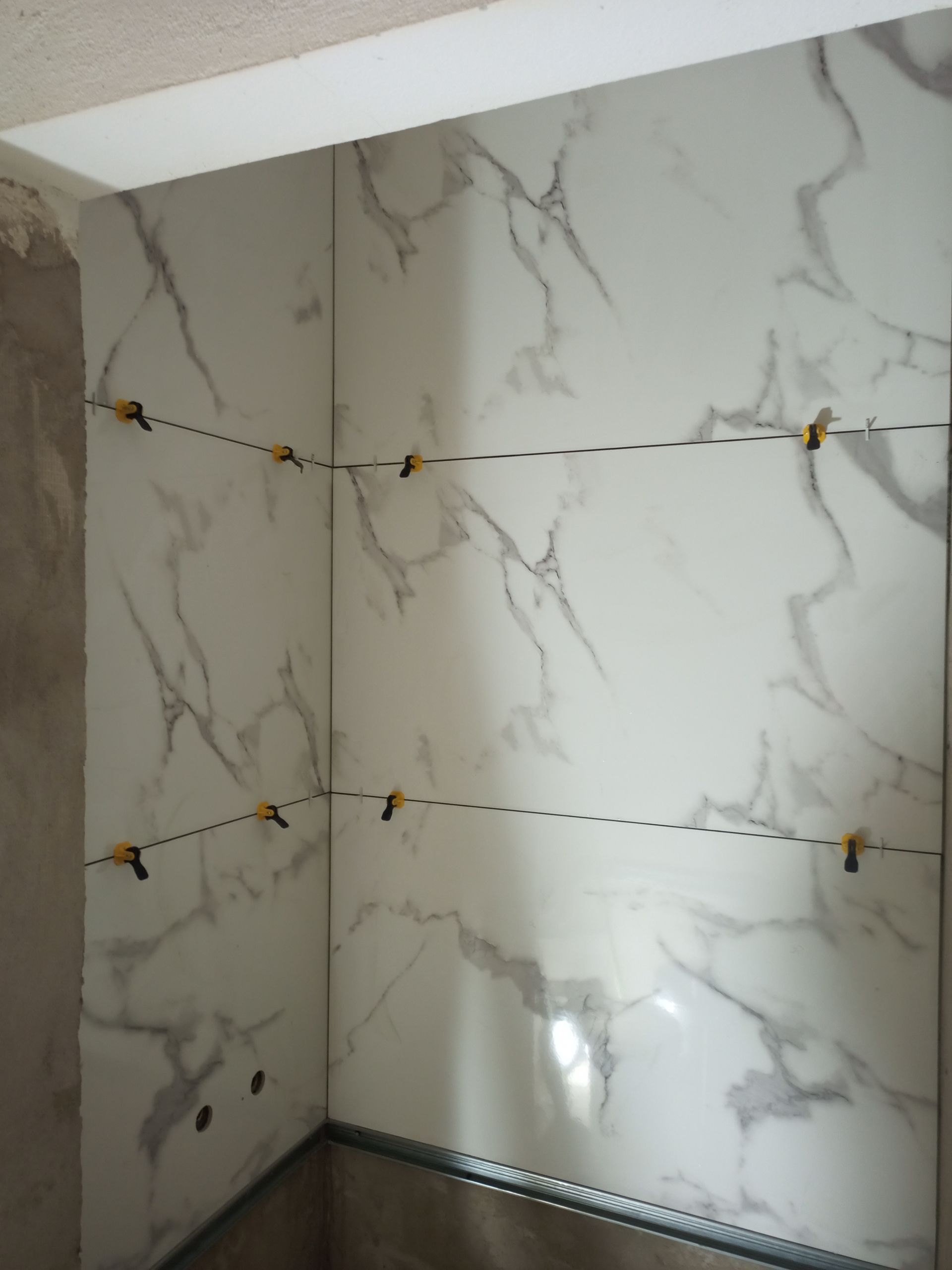 Azulejos brancos com estampa de mármore sendo instalados em um canto da sala, fixados por grampos niveladores.