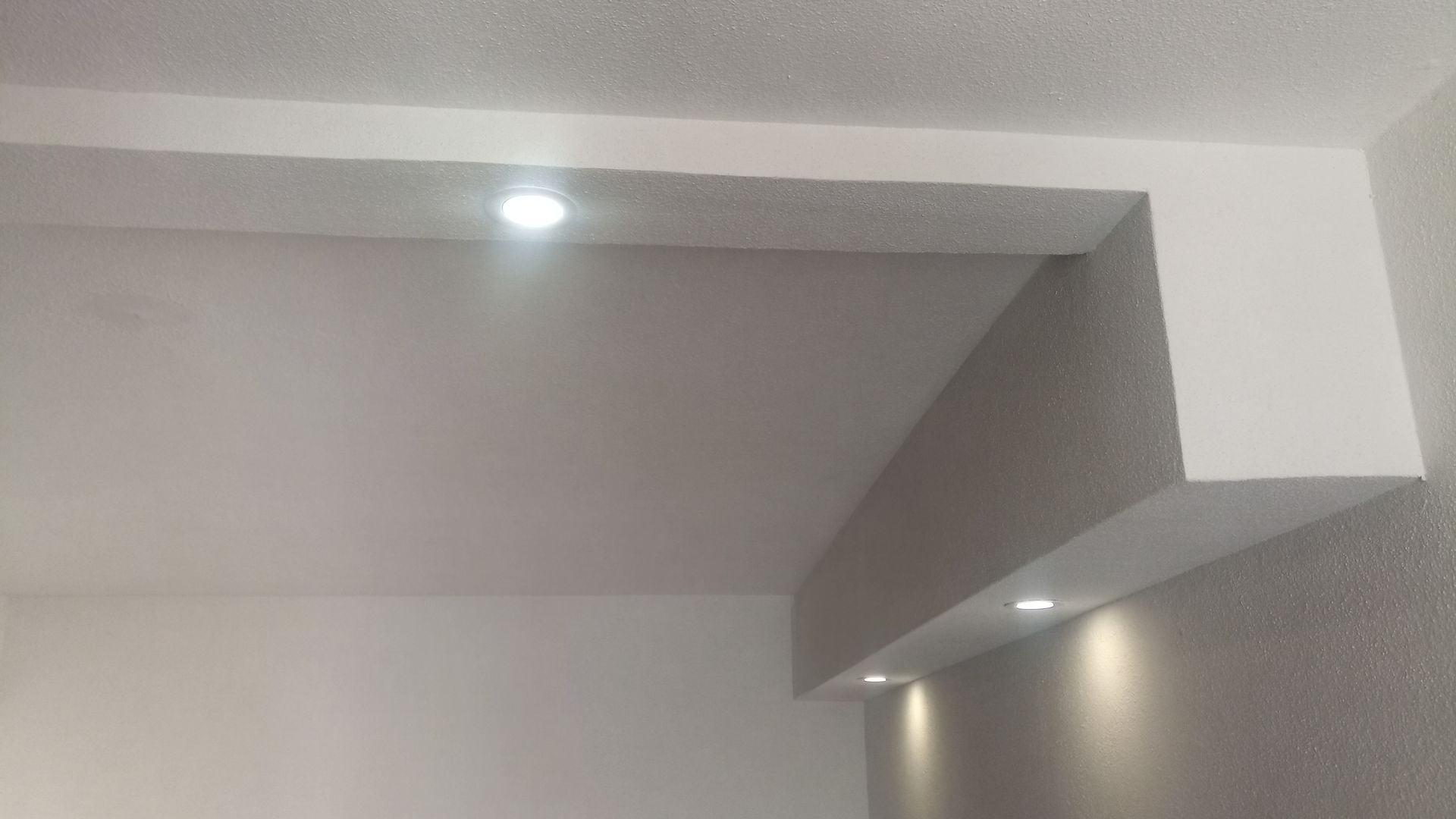 Teto branco com iluminação embutida acima de uma seção de parede cinza e branca.