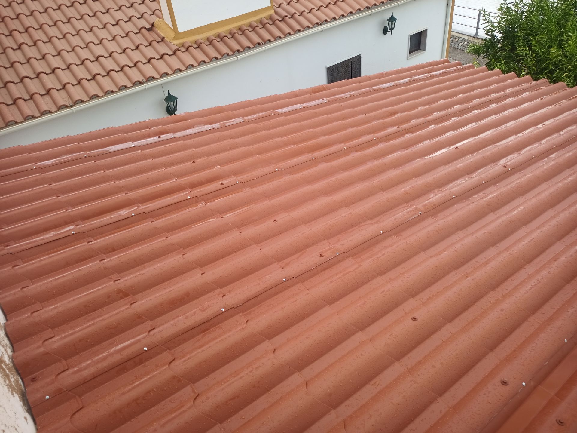 Telhado de metal ondulado vermelho, vista em ângulo. Telhado de telha cerâmica marrom visível ao fundo.