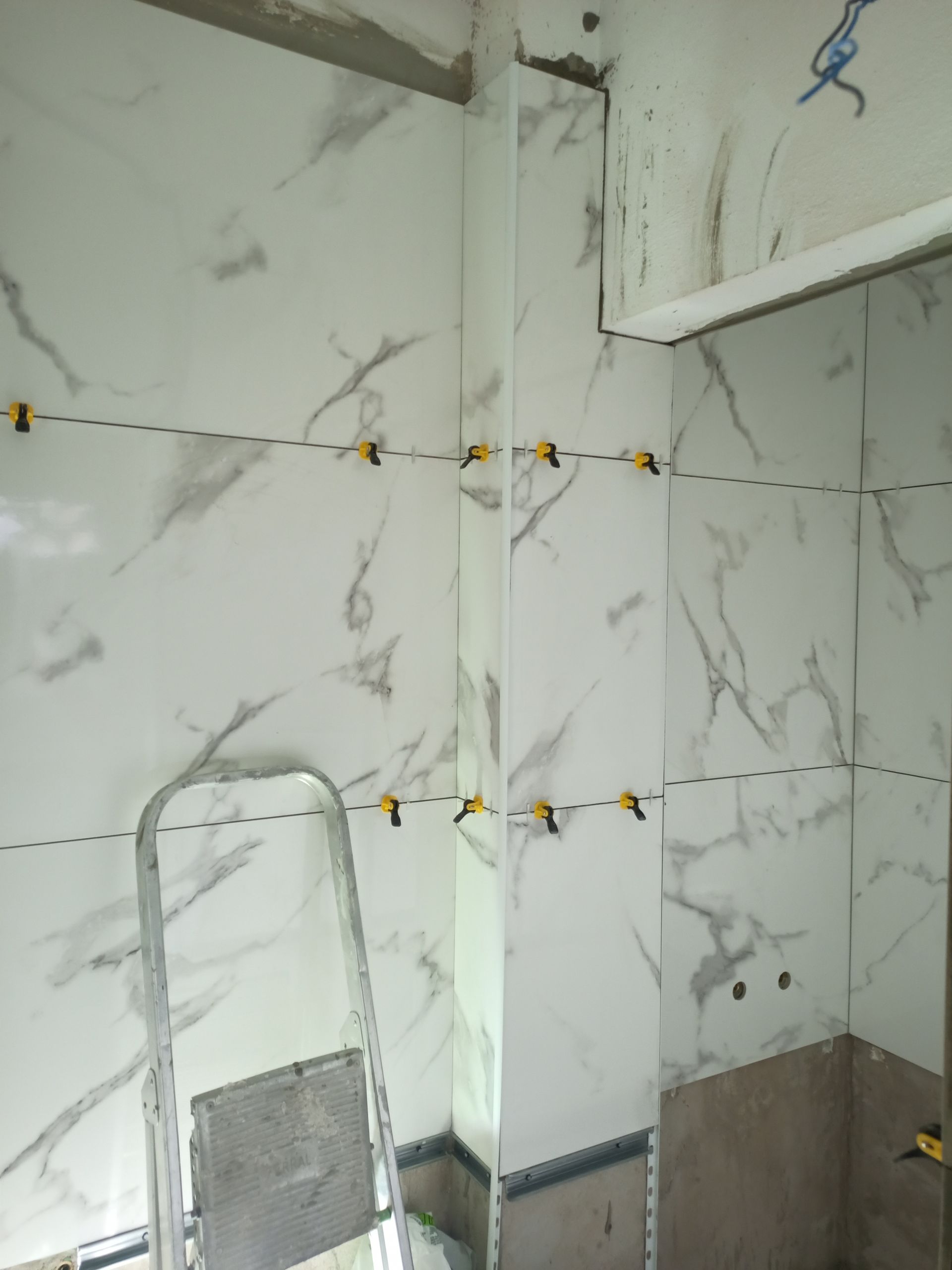 Parede revestida com azulejos em andamento; azulejos brancos com aparência de mármore e grampos de nivelamento. Uma escada de mão está próxima.