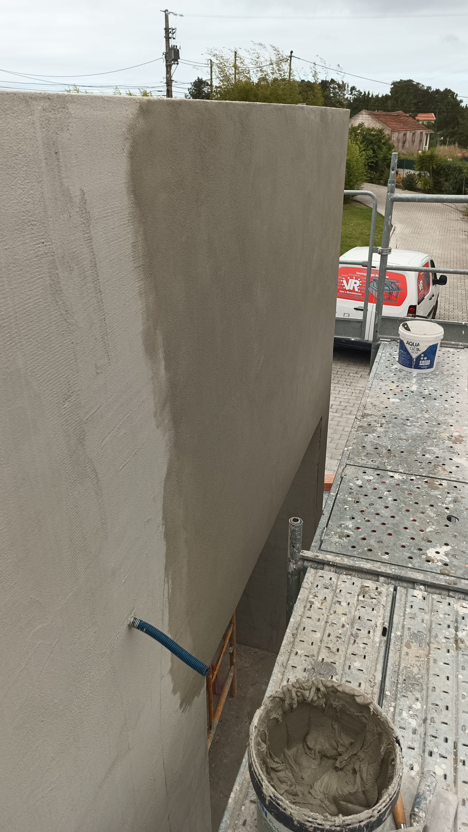 Parede externa com estuque parcialmente aplicado; andaimes e balde de argamassa visíveis.