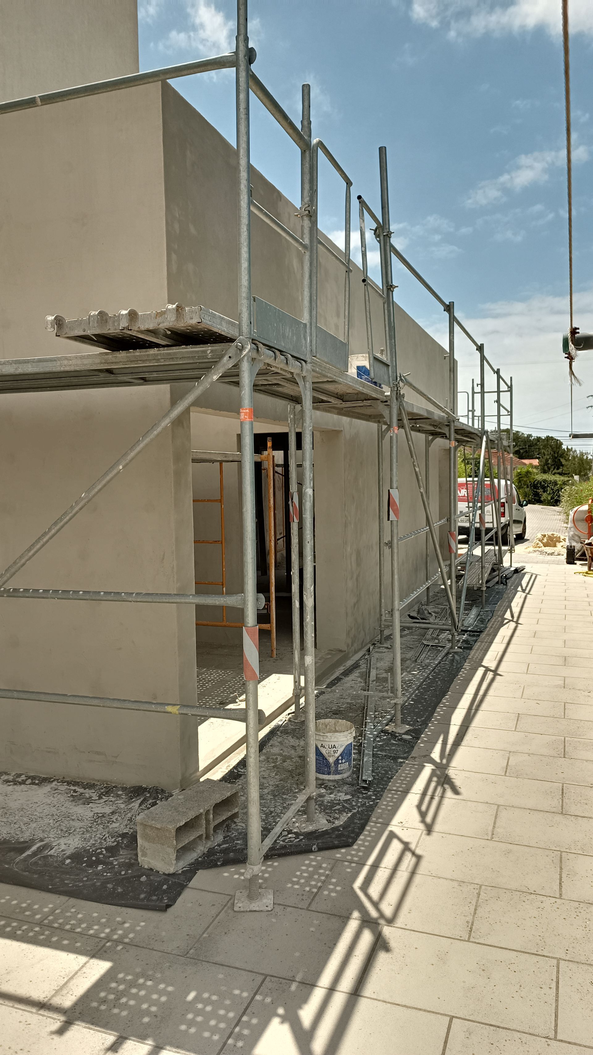 Andaimes na fachada de um prédio em construção; paredes cinza-claro, céu azul e calçada pavimentada.