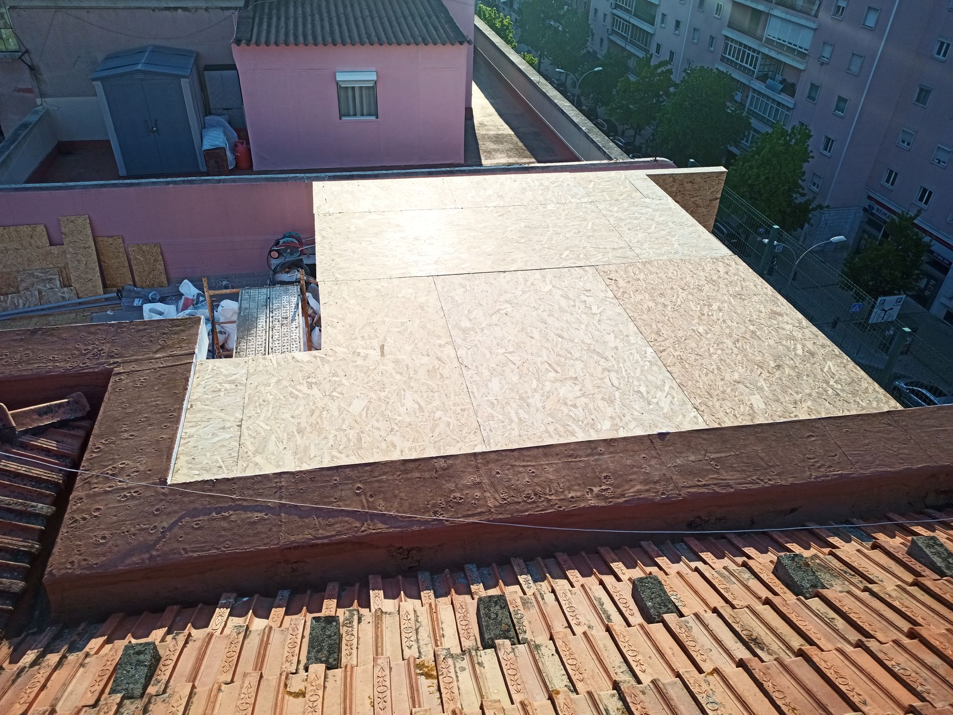 Telhado com painéis de madeira novos. Prédios ao fundo. Dia ensolarado e luminoso.