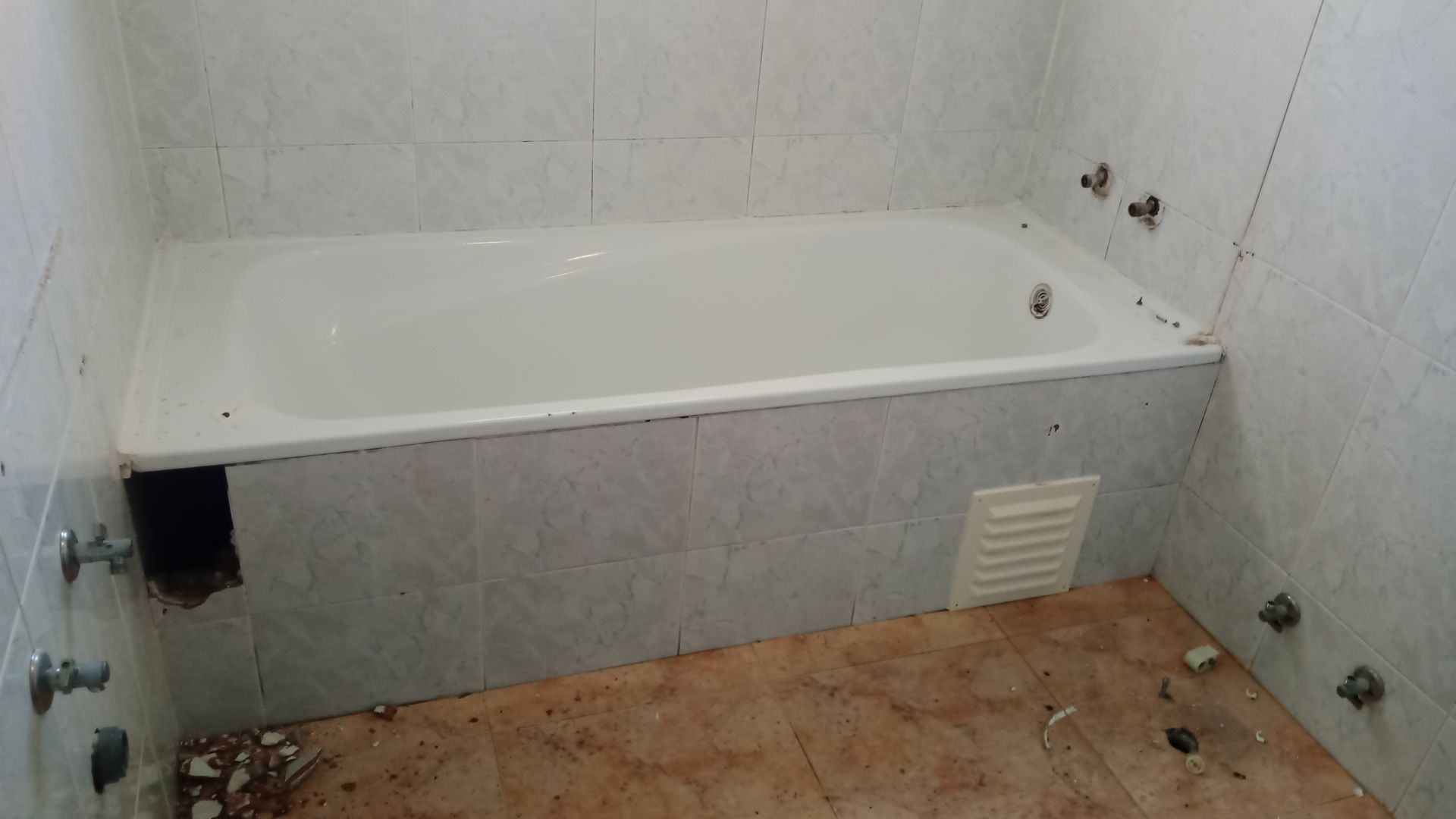 Banheiro com banheira branca, azulejos e acessórios de encanamento. Presença de detritos no chão.