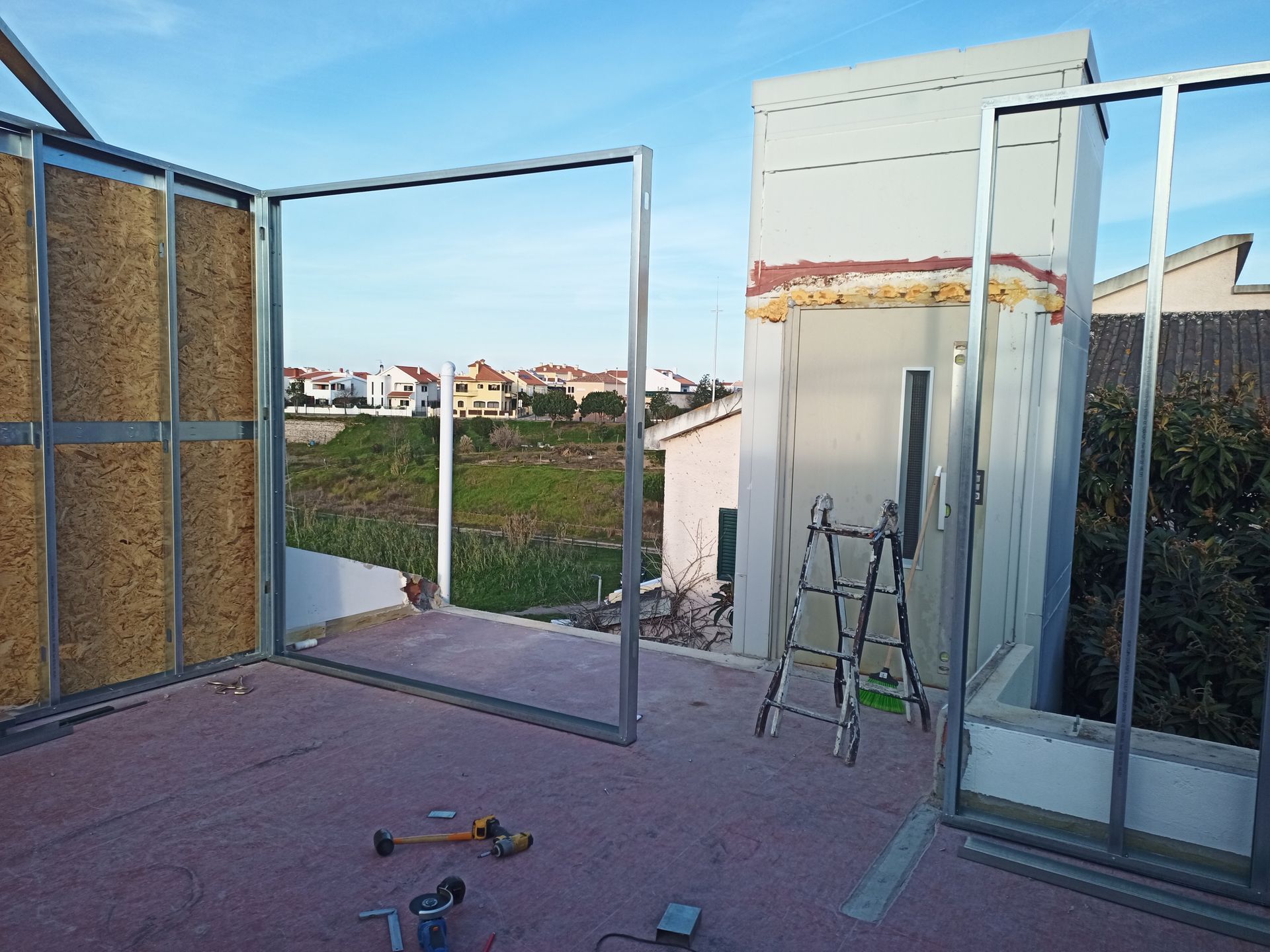 Canteiro de obras. Estrutura metálica para paredes e abertura de porta em um telhado com vista para casas e campos.