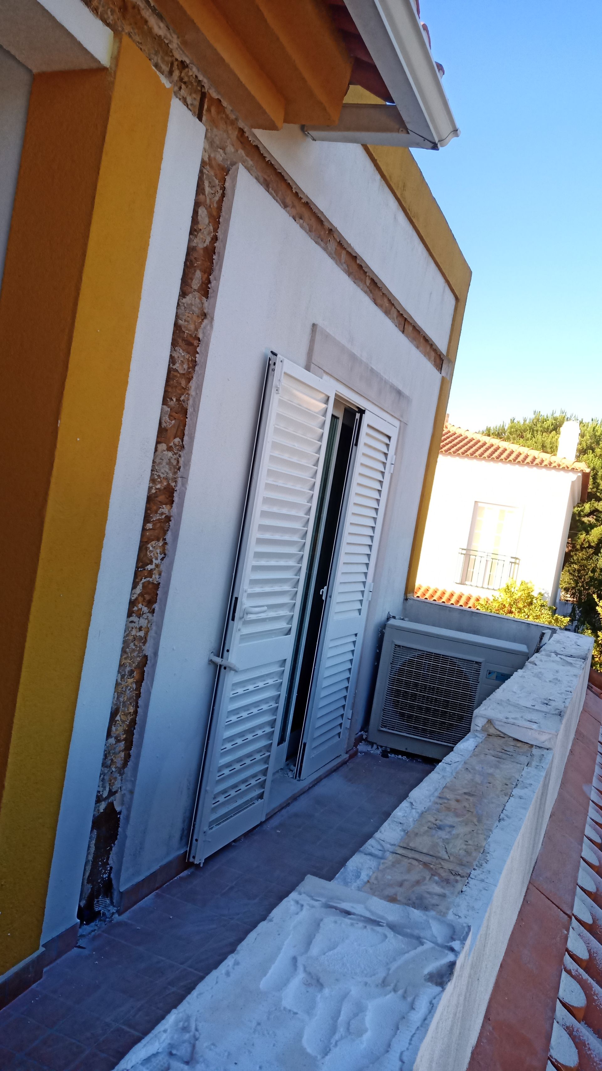 Fachada do edifício com parede amarela, persianas brancas e varanda com parapeito de pedra.