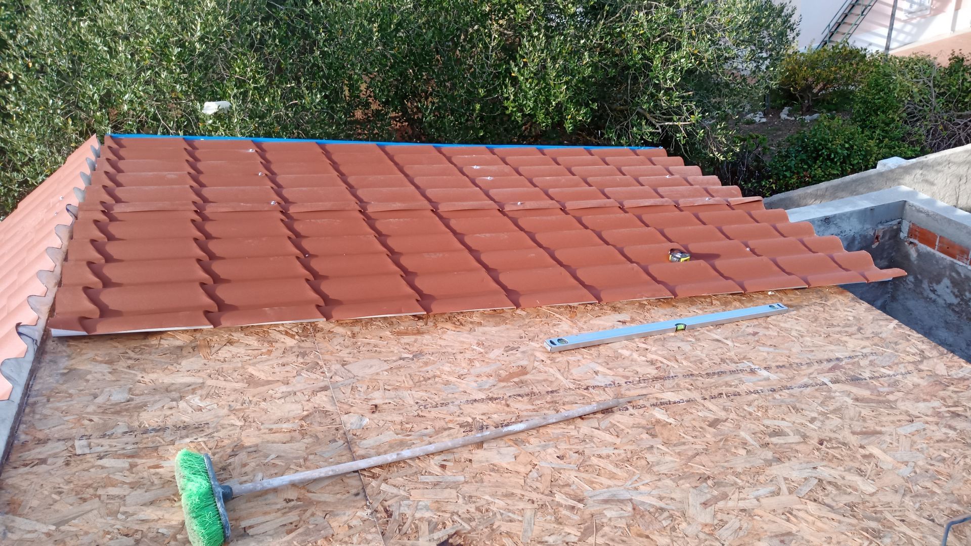 Telhado de telhas vermelhas parcialmente instalado sobre uma base de madeira compensada, com uma ferramenta de cabo verde apoiada sobre a madeira compensada.