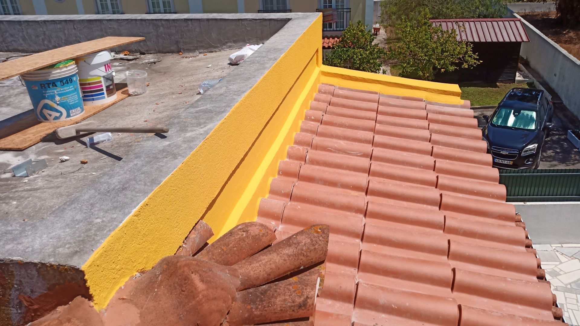 Telhado com telhas laranja e isolamento amarelo, ao lado de uma seção de concreto com latas de tinta e tábuas de madeira.