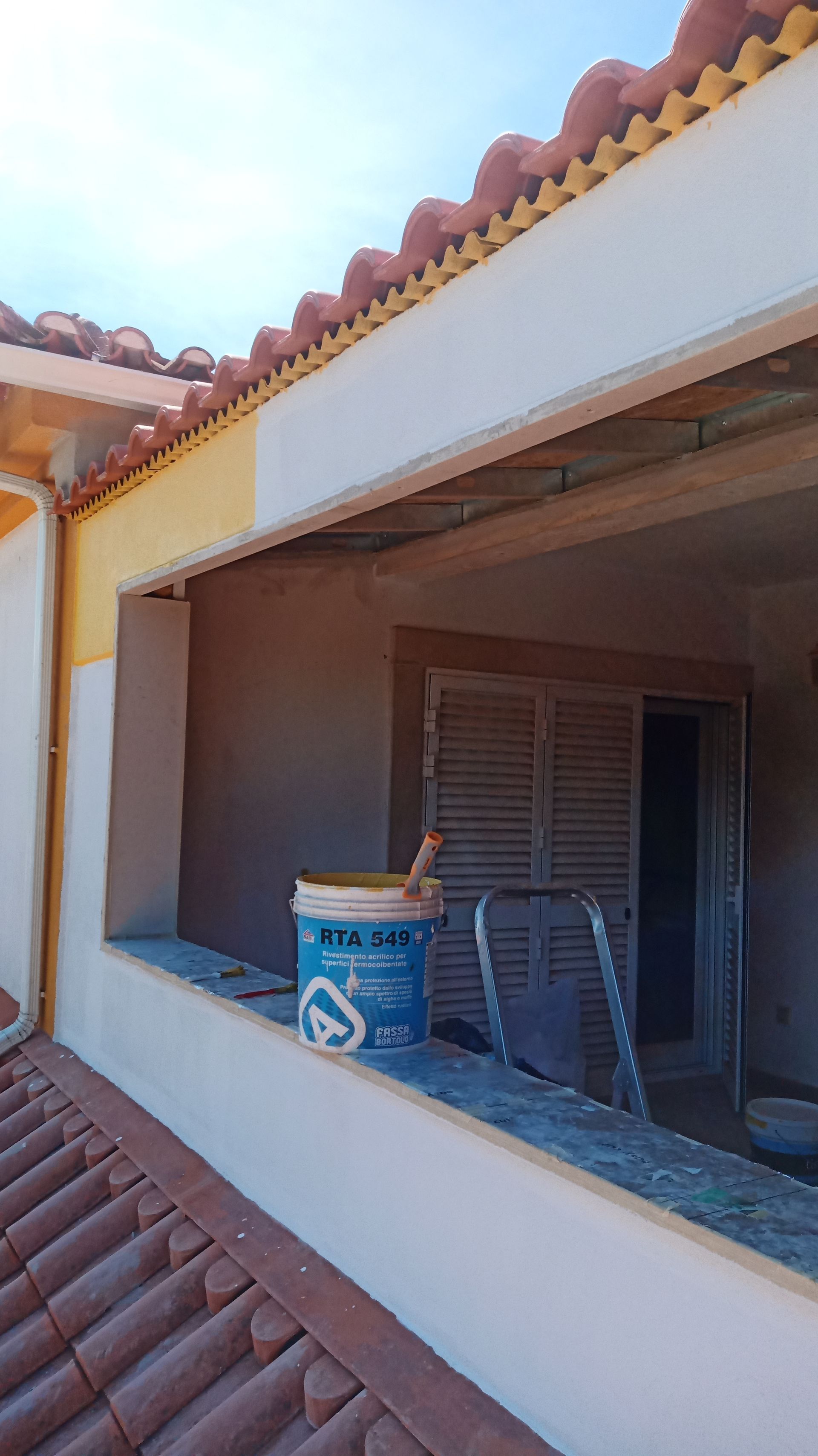 Exterior de um prédio em reforma com a fachada parcialmente pintada de branco e amarelo; um balde de tinta está sobre uma saliência.