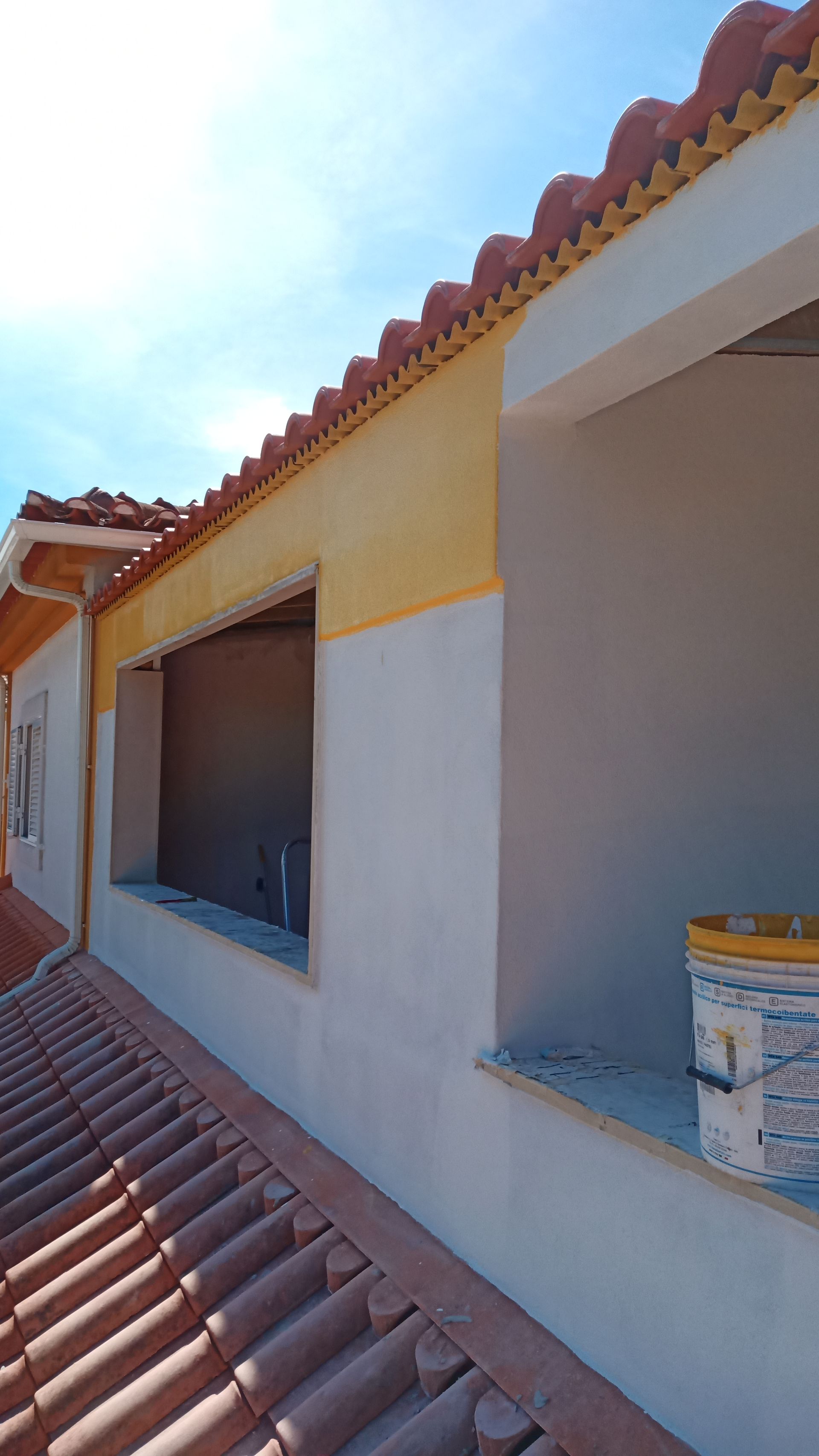 Vista exterior de um edifício com telhado de telhas vermelhas, detalhes em amarelo e paredes brancas; uma janela está tapada com tábuas.