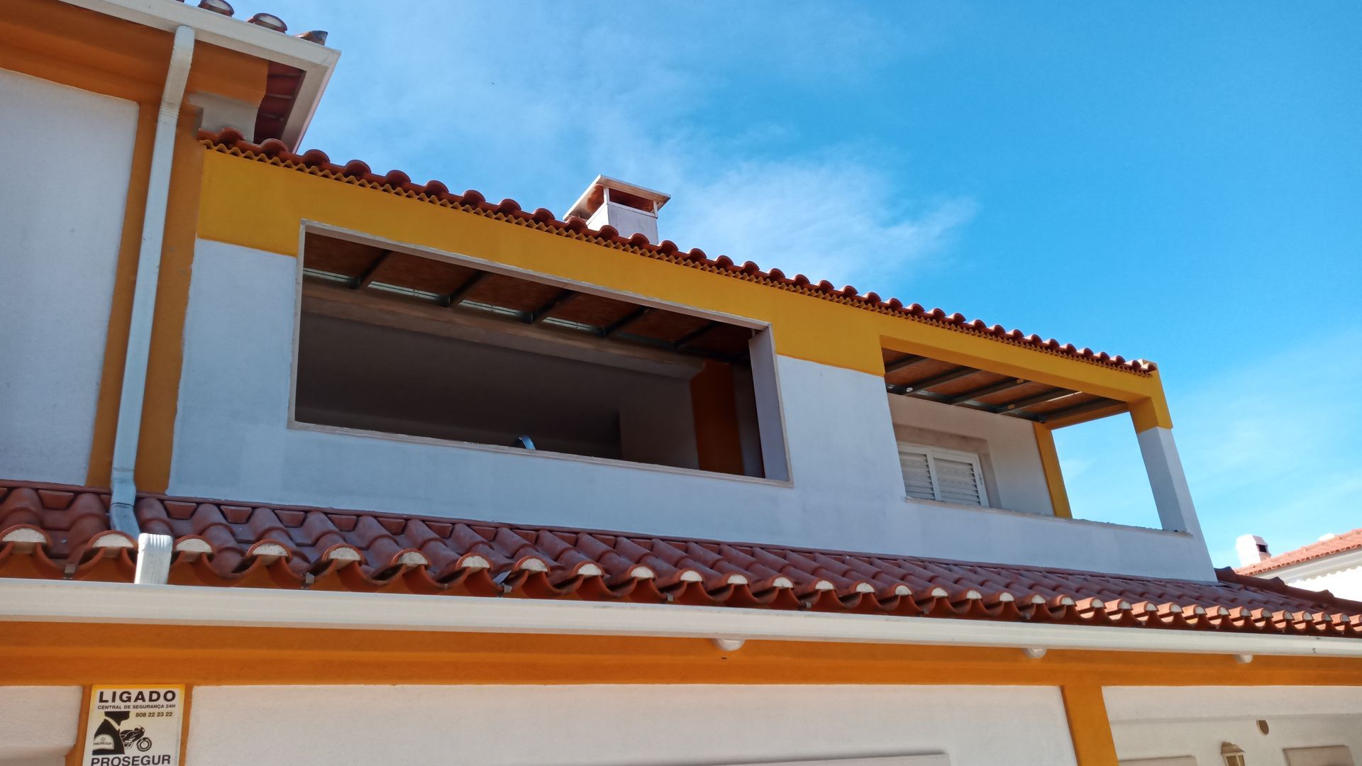 Prédio de dois andares com pintura amarela e branca, telhado de telhas vermelhas e uma grande janela aberta sob um céu azul.