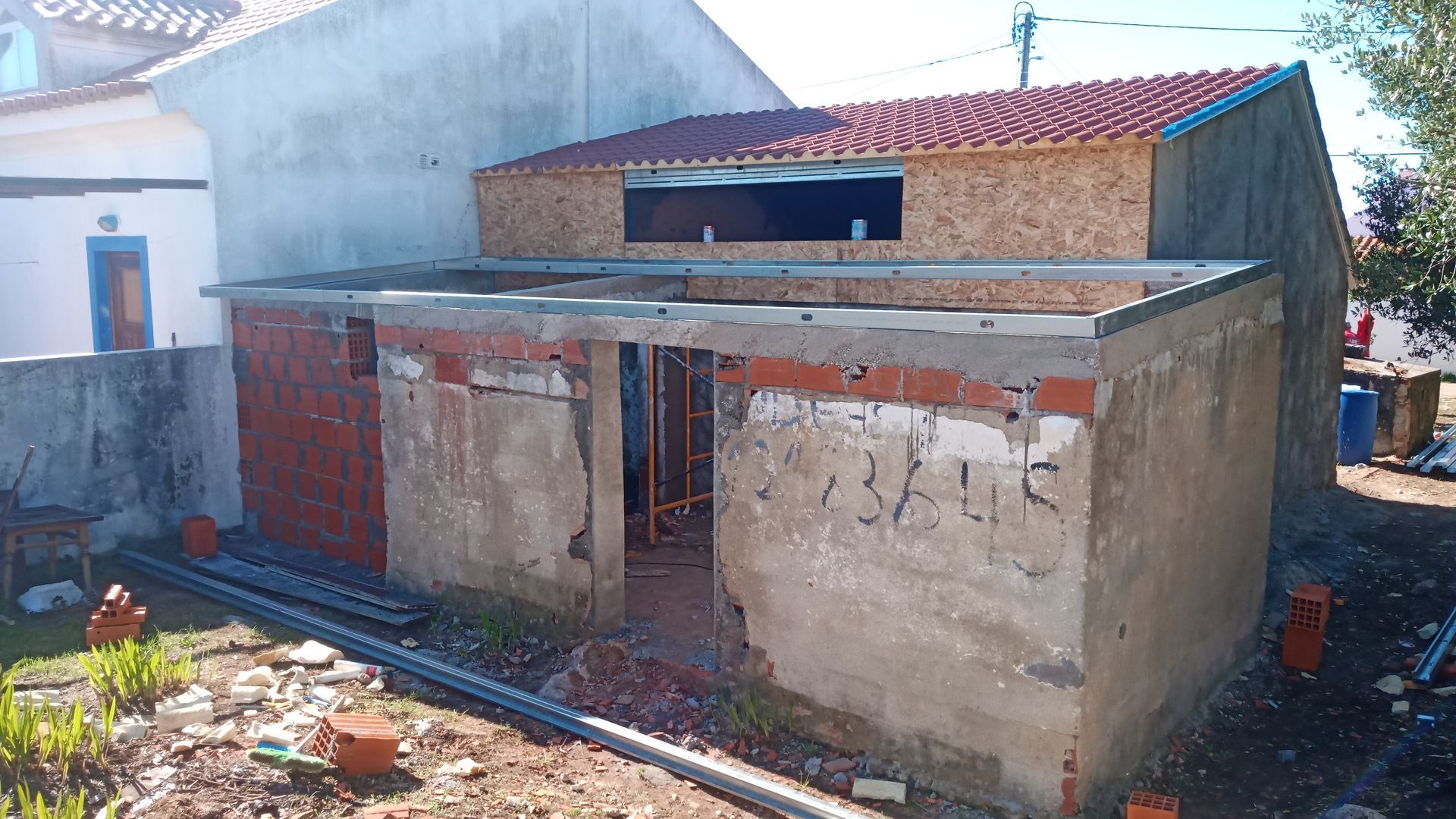 Prédio em construção: estrutura de concreto com tijolos aparentes, um anexo de madeira parcialmente construído e telhas vermelhas.