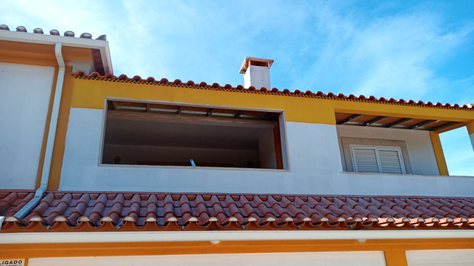 Casa de dois andares com fachada amarela e branca, telhado de telha vermelha e grande janela aberta. Céu azul brilhante.