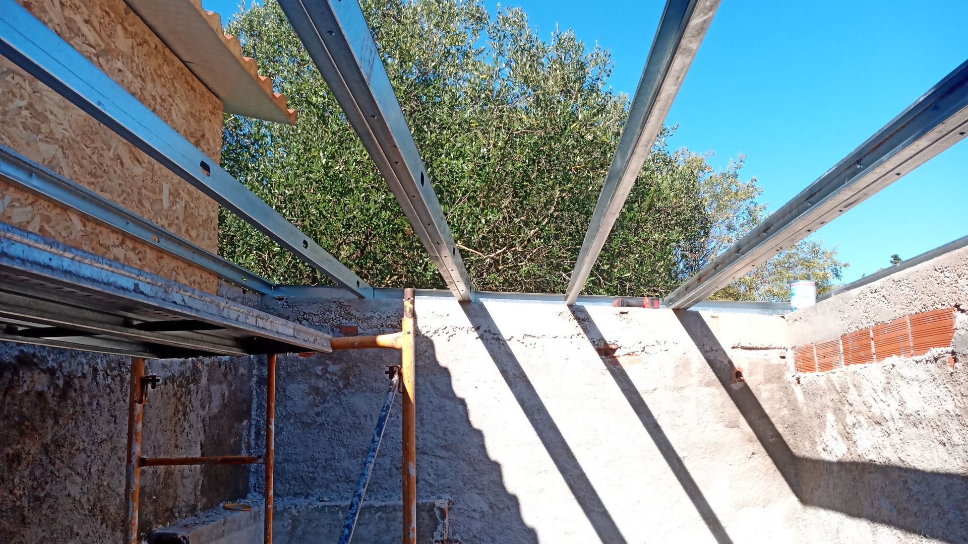 Vigas metálicas instaladas em uma estrutura de concreto e tijolos, tendo como pano de fundo a folhagem e o céu azul.