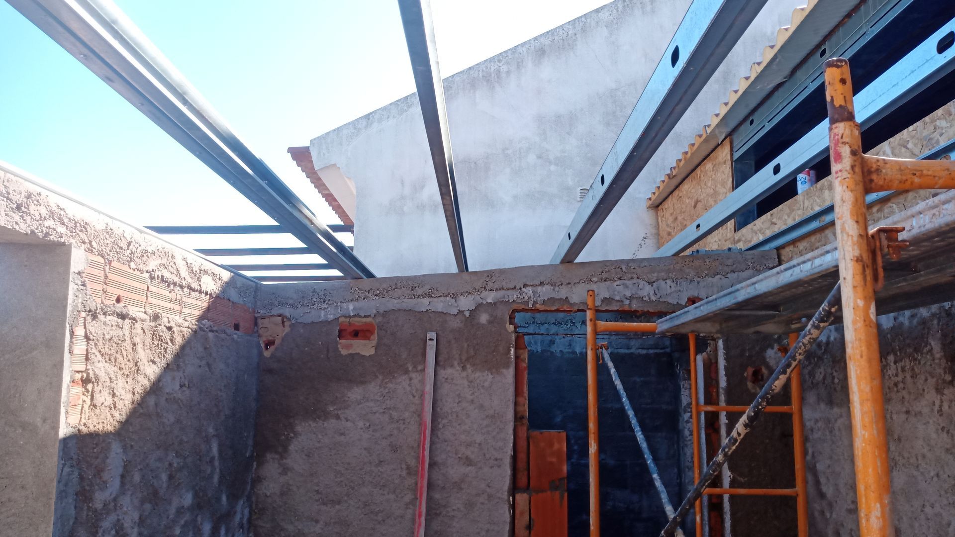 Canteiro de obras com paredes de tijolos aparentes, vigas de metal, andaimes e um céu azul límpido.